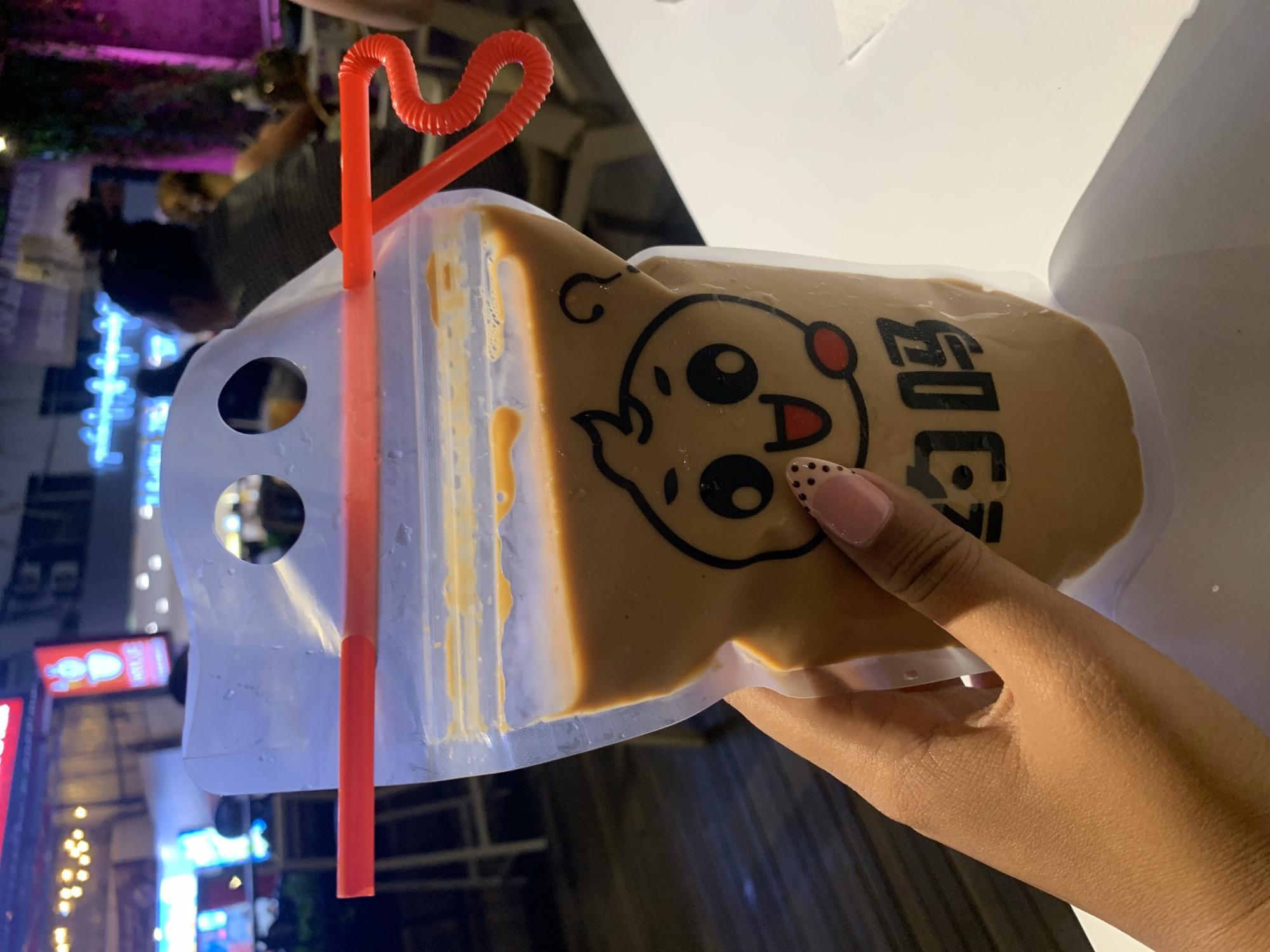 A person's hand with a patterned nail is holding a clear plastic pouch filled with a light brown liquid, possibly a beverage. A red, coiled straw is inserted through the pouch's top opening, with one end inside the pouch and the other extending out. The pouch has a cartoon-like face printed on it, with two circular eyes, a smiling mouth, and rosy cheeks. Below the face are two black Chinese characters. The background appears to be an outdoor setting at night, with blurred lights and structures suggesting a marketplace or street food area in Denpasar, Indonesia. A white table is visible in the foreground, partially obscured by the pouch.