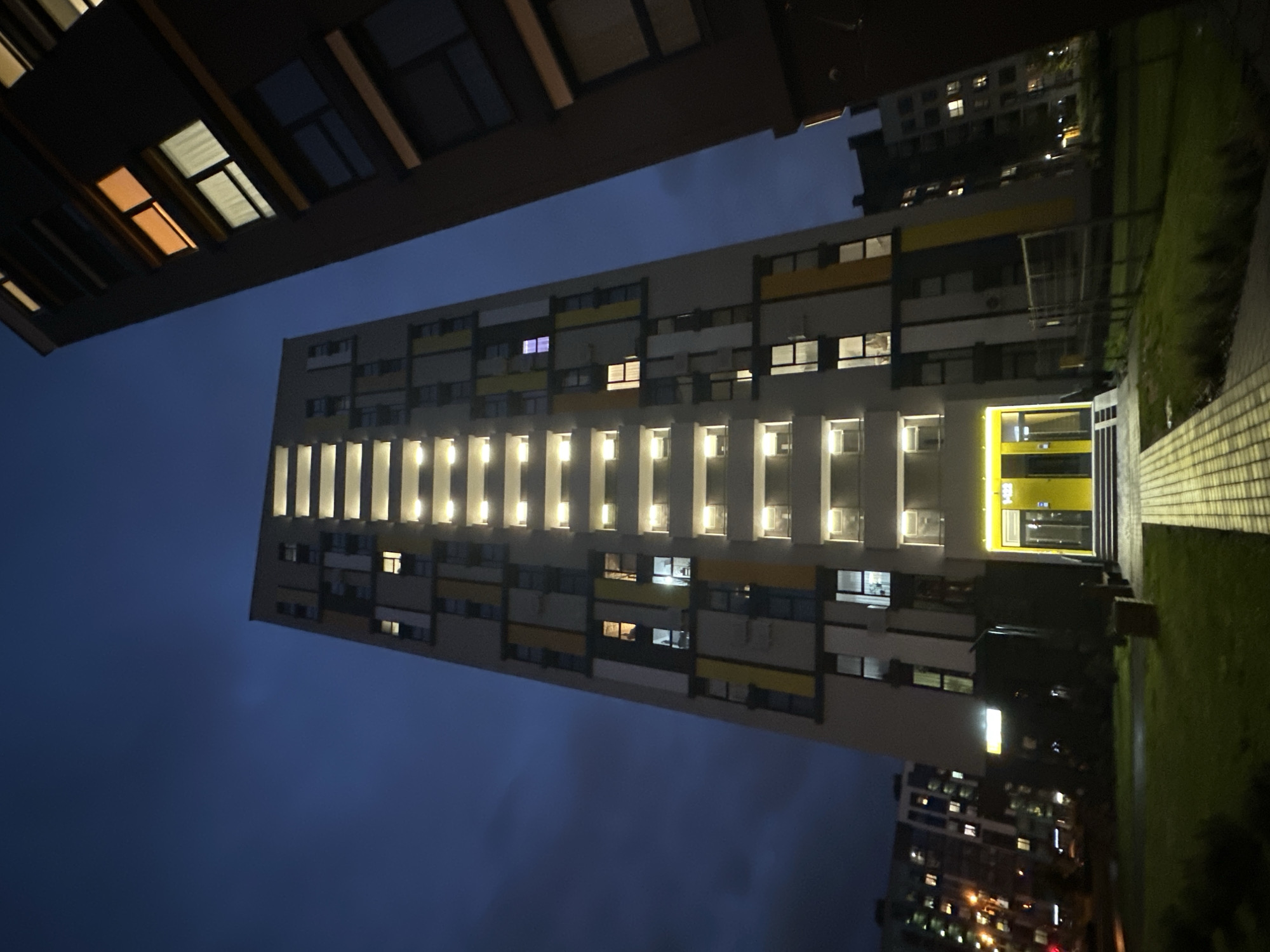 The image depicts a tall, modern apartment building in Бараўлянскі сельскі Савет, Belarus, taken from a low angle at night. The building's facade is primarily white with geometric patterns of black and yellow. Rows of windows are visible, some illuminated from within, creating vertical lines of light. A bright yellow entrance area is also visible. In the foreground, the edge of another building and some greenery are present, partially obscuring the view. The sky is a dark blue, indicating the late hour. Other buildings with illuminated windows are visible in the background. The scene suggests a quiet, residential area at night.