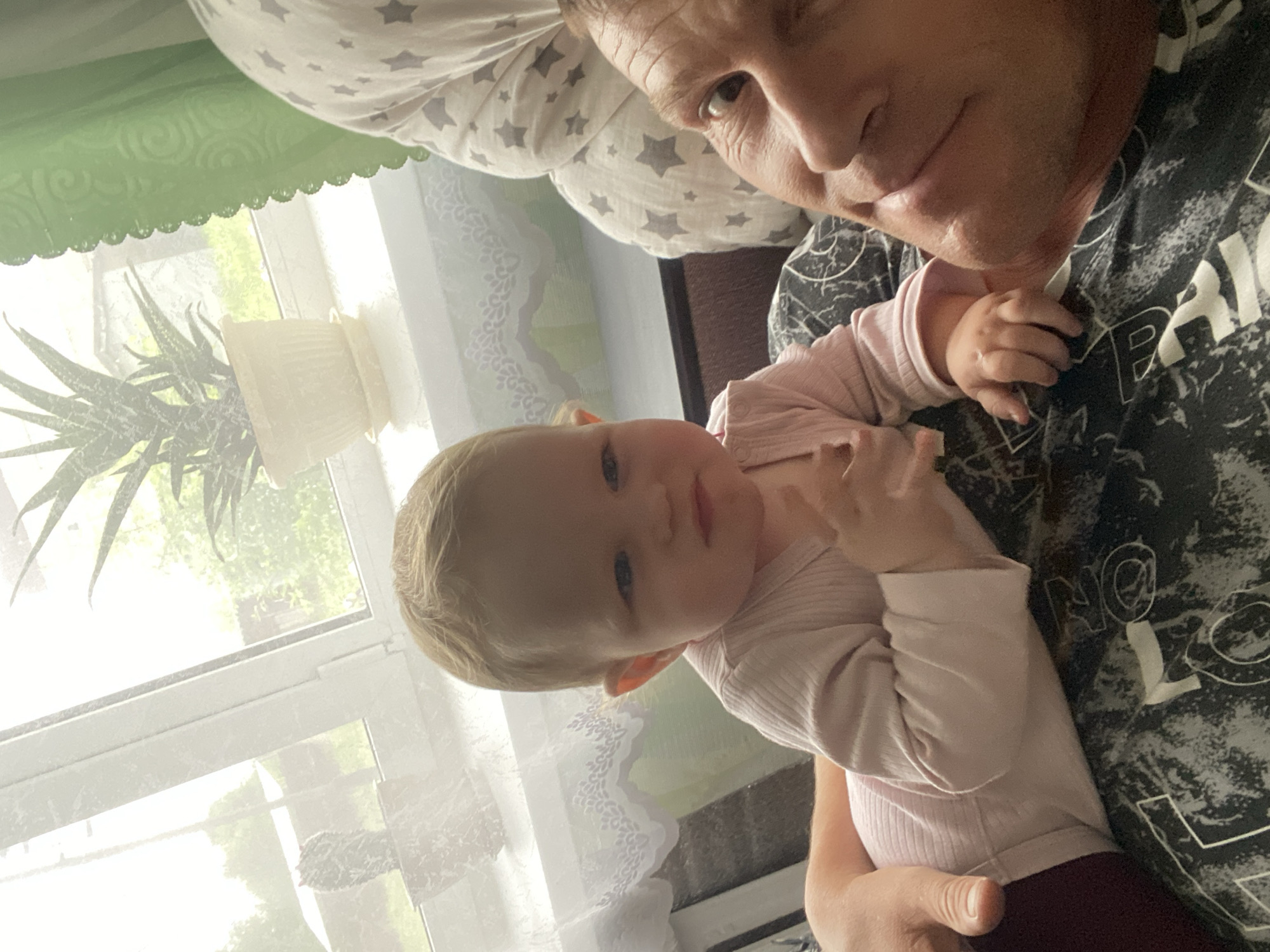 An overhead, vertically oriented photograph captures an intimate indoor scene in Fokino, Russia, featuring an adult and a baby. The image is taken from a high angle, looking down at the subjects.
The main subjects are an adult man and a young baby. The man, with short hair and visible facial lines, looks towards the camera with a gentle expression. He is wearing a dark t-shirt adorned with abstract white patterns and faint, blocky letter-like shapes (e.g., 'P', 'E', 'B') that are not fully legible. The baby, with light blonde hair and blue eyes, is held by the man and also looks towards the camera, appearing calm and content, perhaps with a slight, curious expression. The baby wears a light pink, long-sleeved ribbed top and dark-colored pants or leggings, with their small hands resting near the adult's chest.
The activity depicts a moment of affectionate holding or bonding between the adult and the baby. The setting appears to be a brightly lit room, likely a home, near a window. Through the window, lush green foliage is visible, suggesting a daytime scene with natural light. On the windowsill, a light-colored potted plant with spiky leaves, possibly an aloe vera, is clearly visible. A decorative green curtain with a scalloped edge hangs above the window. Behind the adult's head, a light-colored pillow patterned with grey stars is partially visible. The overall mood of the image is warm and tender, highlighting a loving interaction.