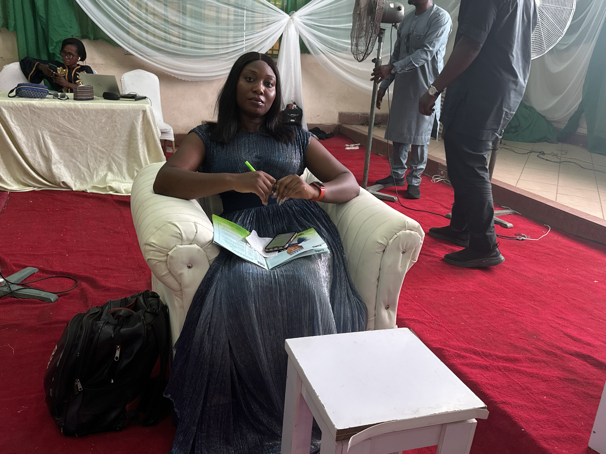 A woman is seated in a cushioned armchair on a red carpet in an indoor space. She is wearing a dark, possibly navy, dress with a shimmering texture, and is holding a green pen in her hands. A booklet and a smartphone rest on her lap. Next to her is a small, white, wooden table. To the left, on the carpet, there is a black backpack. In the background, there are two men standing near a fan and a woman seated at a table with a laptop. The setting appears to be some sort of event, possibly in the city of Unknown, Unknown, decorated with white and green fabrics.