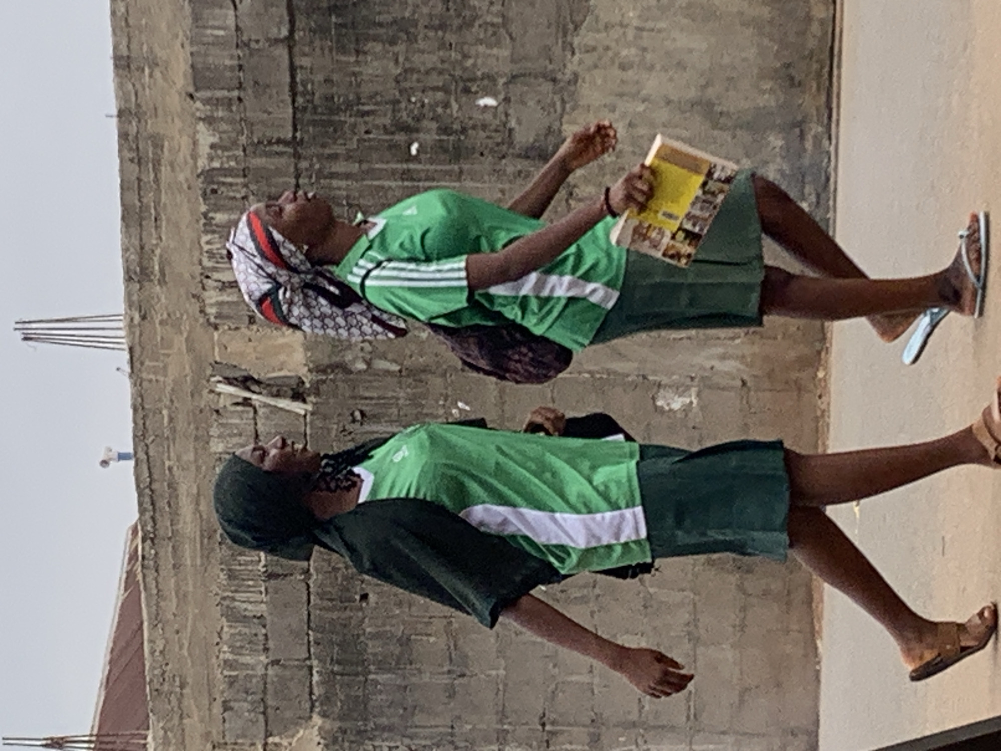Two young women are walking along a paved area adjacent to a rough, unfinished concrete building. The woman on top is wearing a green shirt with white stripes, dark green shorts, and a patterned headscarf, and she is holding a book. She has light-colored sandals on her feet. The woman on the bottom is wearing a green shirt with a white stripe and dark green shorts with a dark-colored head covering. She has brown leather sandals on her feet. They are likely in Akwanga, Nigeria.