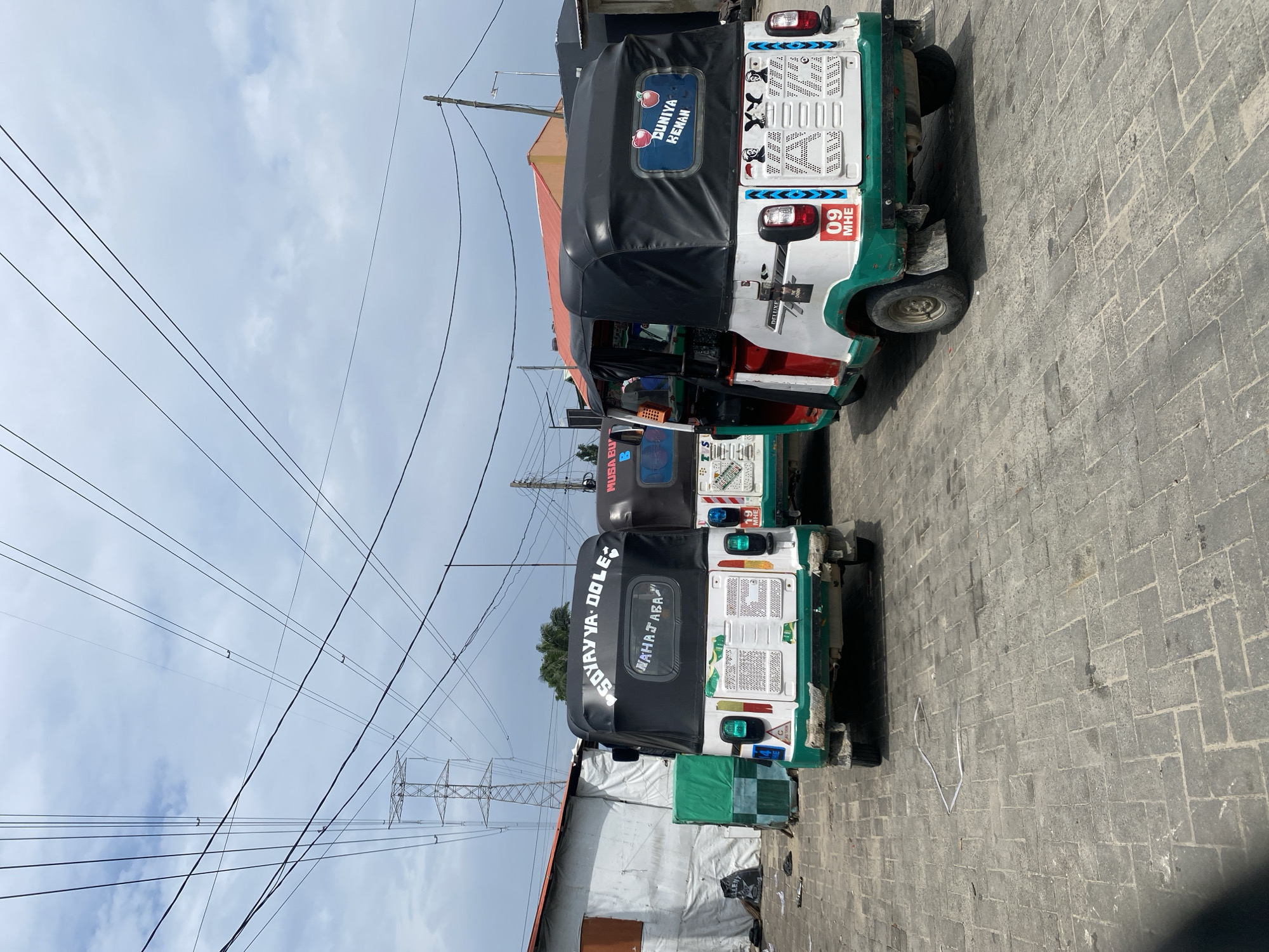 The image shows a street scene in Ado, Nigeria, with three colorful auto-rickshaws, locally known as "keke naphop," parked on a paved surface. The auto-rickshaws are white with green and red accents, and each has a black canopy. The first auto-rickshaw on the left has the inscription "SOYAYYA DOLE" and "WAHAJABA" on its back. The second auto-rickshaw in the middle is partially visible and has "MUSA BU" written on it. The third auto-rickshaw on the right displays "DUNIYA KENAN" along with two red apples on its rear. The auto-rickshaws are facing away from the camera, with the one on the right having its door open. In the background, there are buildings, including a white structure with a corrugated iron roof and a red-tiled roof structure. Overhead, a network of power lines and a large electricity pylon are visible against a cloudy sky. The ground is covered with gray interlocking paving stones. There are no people visible in the frame. The scene appears to be a commercial area or a parking lot.