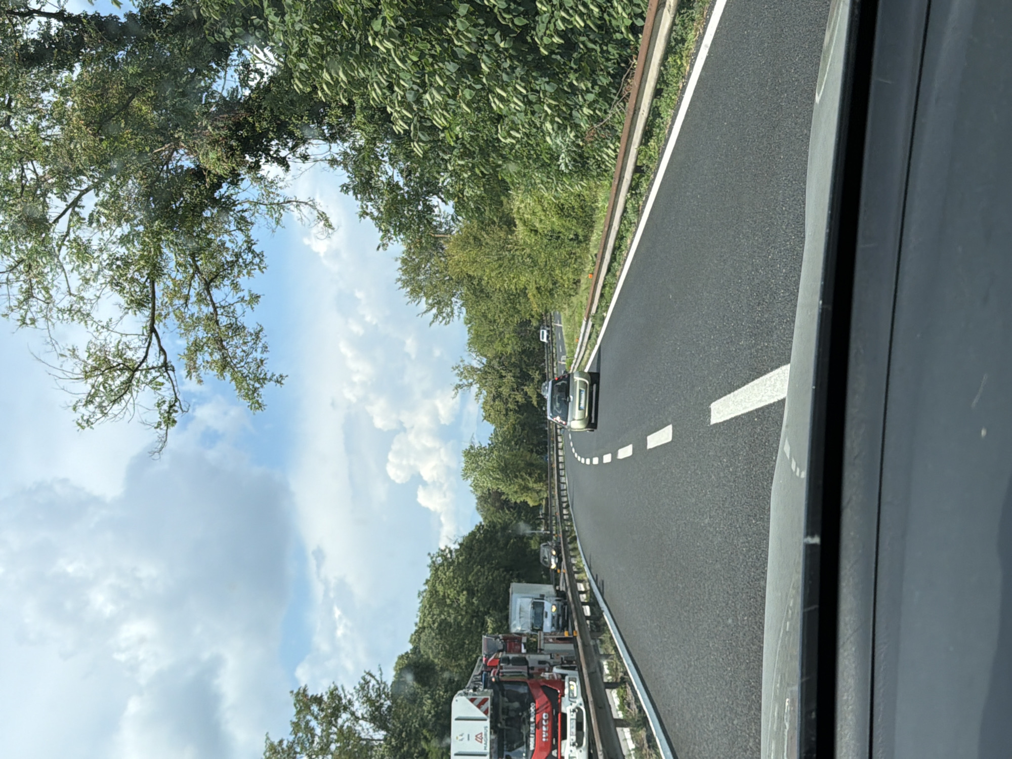 The image shows a road in Basaldella / Basandiele, Italy. The road is paved and has white lane markings. There are several vehicles on the road, including cars and trucks. Some of the trucks appear to be emergency vehicles, with one red and white truck that looks like a fire engine. The scene is bordered by trees and foliage on the left and a guardrail on the right. The sky is partly cloudy. The overall impression is of a traffic jam or a stopped procession of vehicles, possibly due to an emergency. The time of day appears to be daytime, and the weather is fair. No people are clearly visible, but the presence of emergency vehicles suggests a possible event requiring their attendance. Some text is visible on the side of the fire engine, including "MAGIRUS" and "IVECO". There also appears to be some text on the side of another truck that is difficult to read. The image is taken from a vehicle, looking forward along the road.