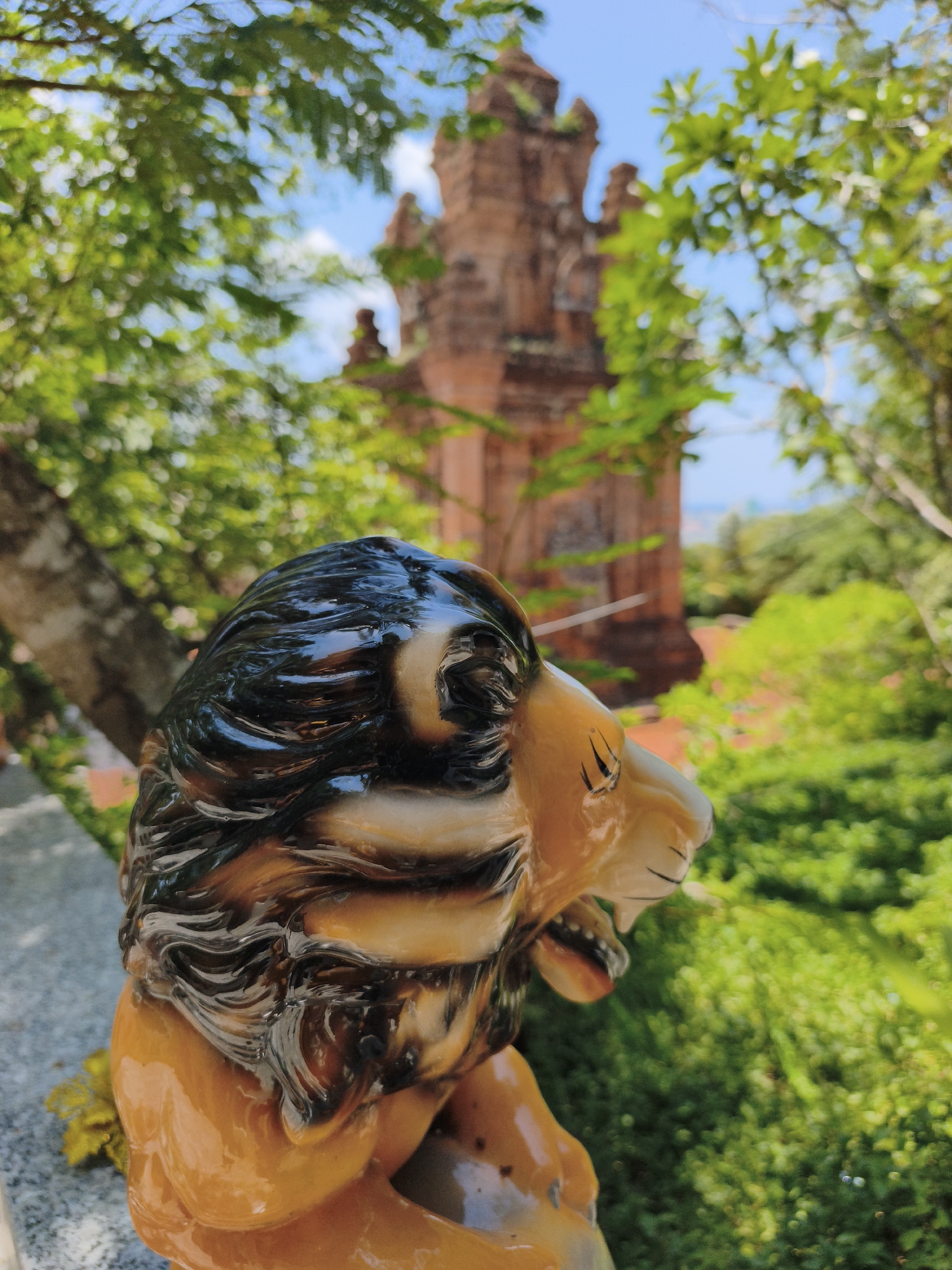 The image shows a ceramic statue of a lion in the foreground, with its head and upper body visible. The lion is depicted with a flowing mane, an open mouth, and a snarling expression. Behind the statue, out of focus, is a brick tower, likely part of a Cham temple complex, and lush green foliage. The sky is blue with some clouds, suggesting a clear, sunny day. This scene is characteristic of the Cham towers found in and around Tuy Hoa, Vietnam, such as the Thap Nhan (Nhan Tower) which is a prominent landmark in the city. The statue appears to be placed on a stone ledge or railing, overlooking a landscape of trees and vegetation.