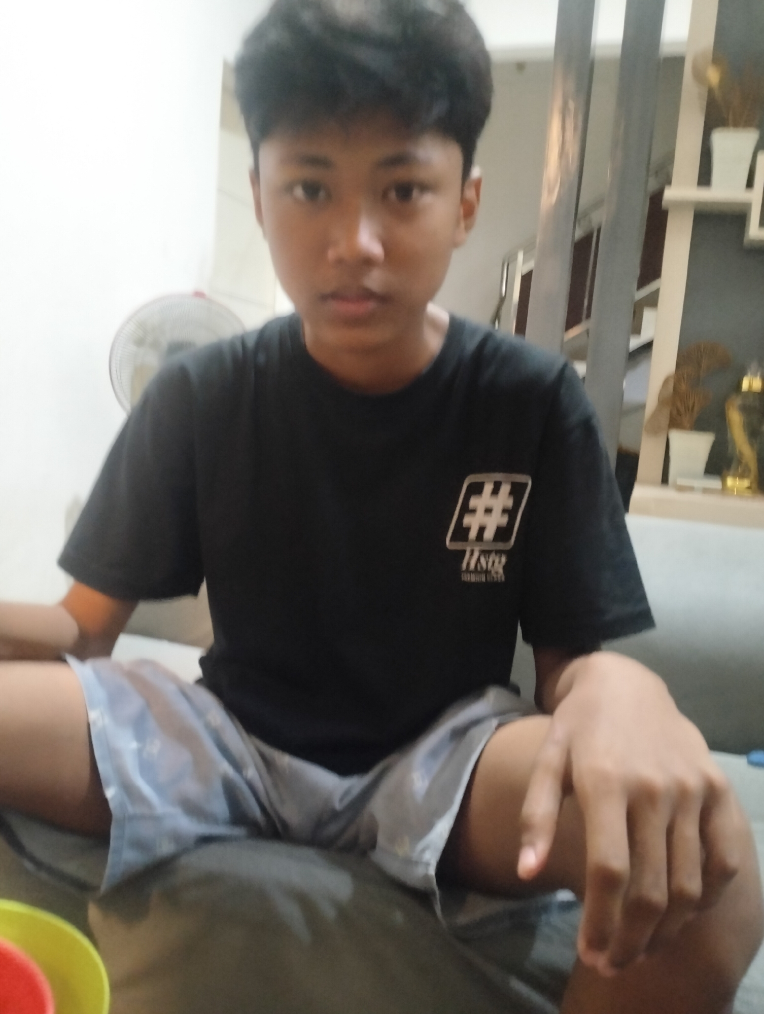 A young male is seated on a couch, looking towards the camera. He is wearing a black t-shirt with a white graphic on the left chest and light blue patterned shorts. His legs are crossed. To the left of the frame, a portion of a red and yellow bowl is visible. Behind him, a standing fan is partially visible. The background includes shelves with decorative items and a staircase. The scene appears to be indoors, within a residential setting in Sidodadi, Indonesia.
