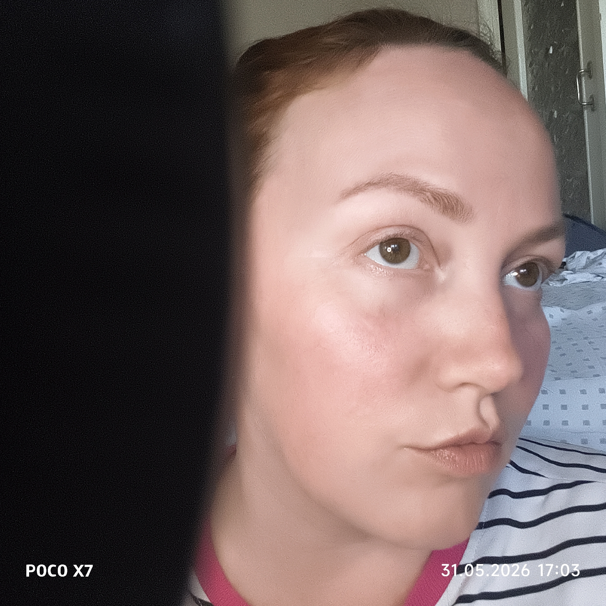 A close-up, low-angle photograph shows a fair-skinned person with brown hair and brown eyes. The person's face is the primary subject, taking up a significant portion of the right side of the frame. The person is wearing a white shirt with black stripes and a pink collar.
On the left side of the image, a large, undefined black object obscures part of the scene. The background is ambiguous but appears to include a light-colored wall and a bed with a patterned sheet.
The image includes text in the lower left corner ("POCO X7") and the lower right corner ("31.05.2026 17:03"). The location and city are unknown.