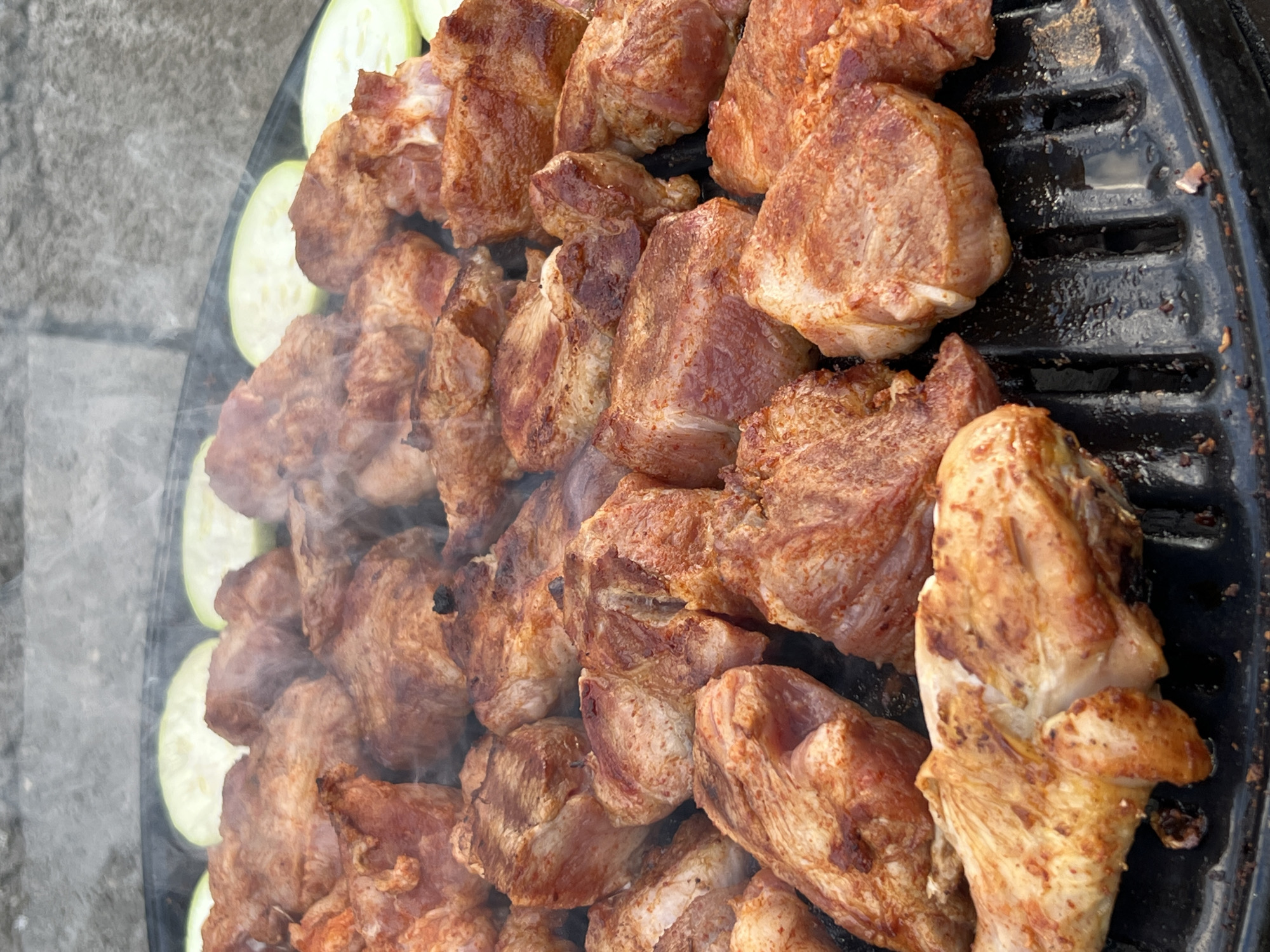 This image depicts food actively cooking on a black, ribbed grill outdoors in the city of Unknown, Unknown. The grill is laden with numerous pieces of browned, marinated meat, primarily cubed pork, alongside at least one golden-brown chicken piece in the foreground. Several light green, round slices of zucchini or squash are positioned along the back edge of the grill. Visible smoke rises from the cooking items. The background shows a textured, light gray surface, consistent with a concrete wall or pavement.
