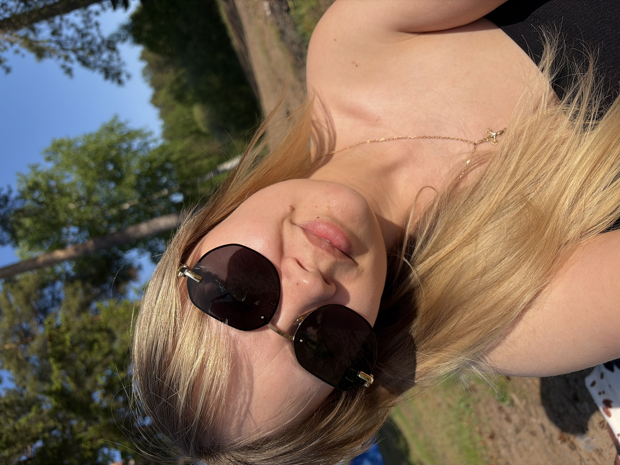 A young woman with fair skin and blond hair is photographed from a low angle, likely outdoors in a wooded area. She wears dark sunglasses and a thin gold necklace. She is dressed in what appears to be a black top, though the full garment is not visible. The background consists of trees and a bright blue sky, suggesting the location is likely a park or natural area. The photo was taken during daylight, with a clear sky, in Chaikovsky, Russia.