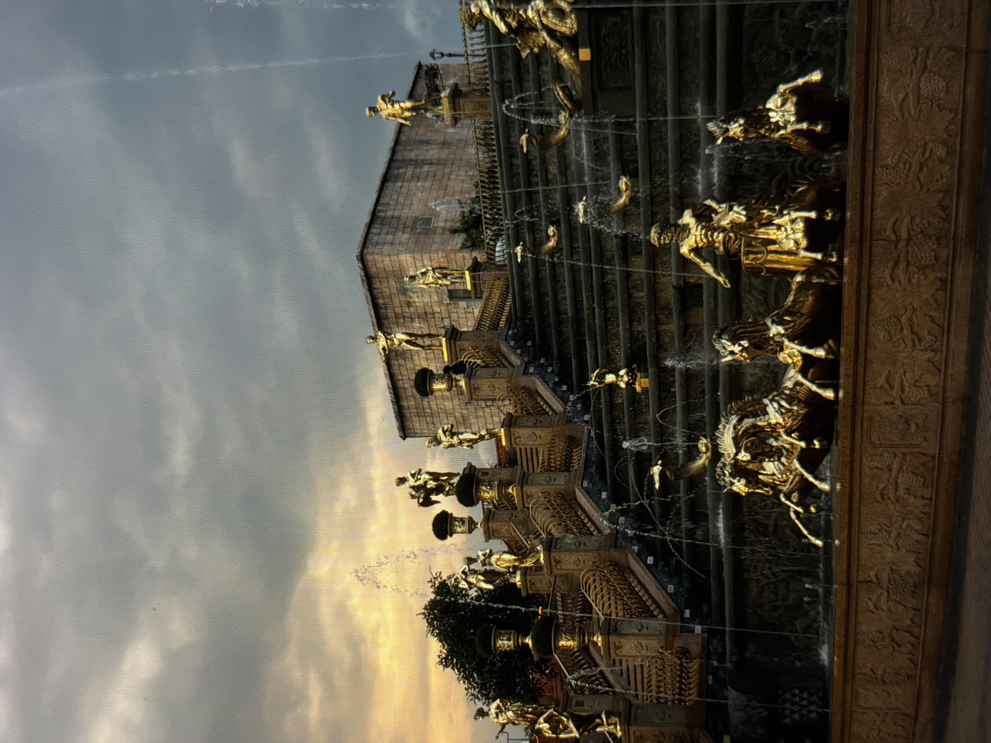 The image depicts the Golden Bridge in Đà Nẵng, Vietnam, viewed from a low angle. The bridge is a multi-tiered structure with golden statues adorning its edges, lined with running water. In the background, a building with brick-like facade is visible. The sky is overcast, with hints of sunset.