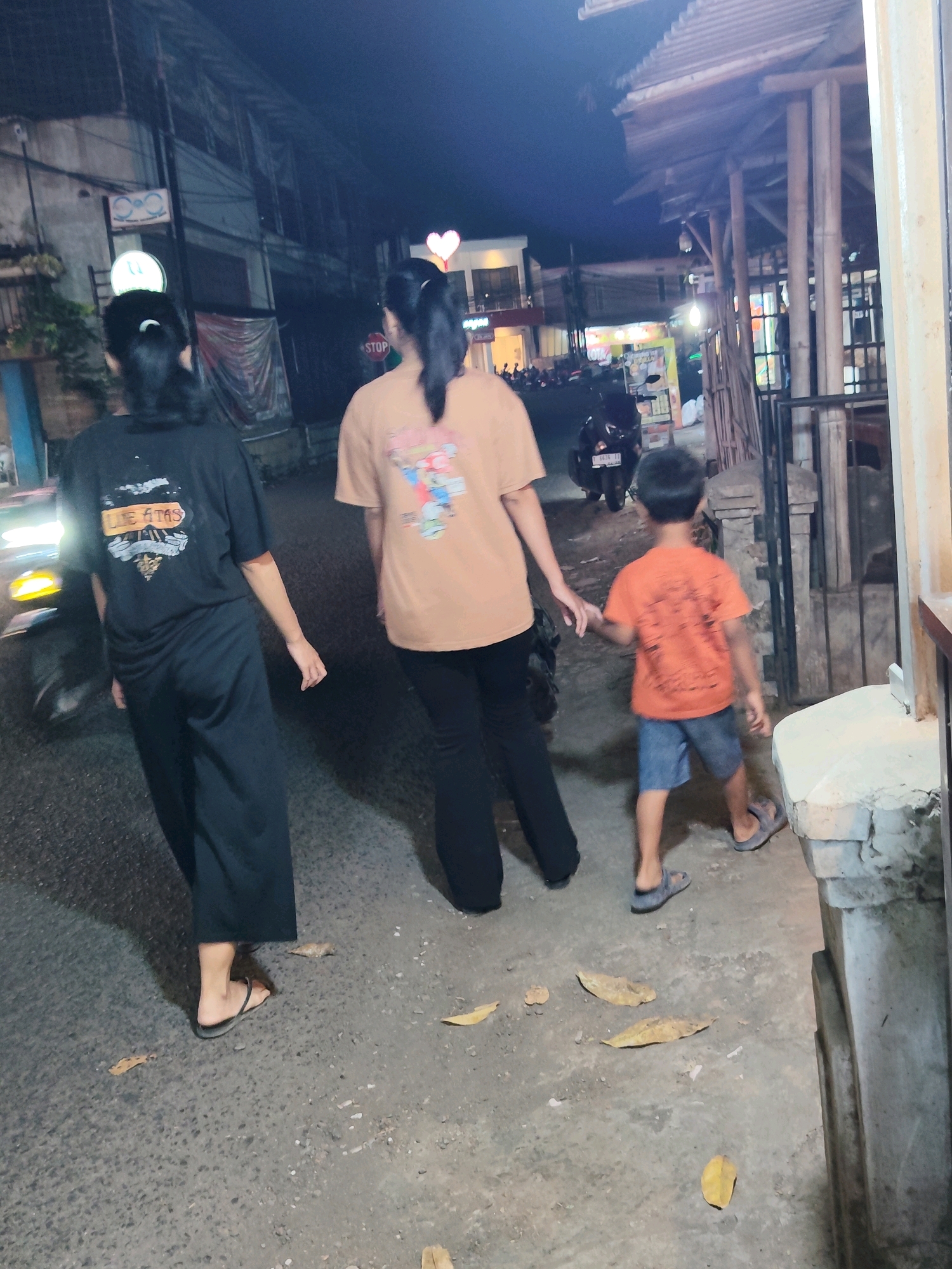 Three people are walking on a paved street at night in Purwakarta, Indonesia. A woman on the left wears a black t-shirt and black pants and is walking away from the camera. To her right, another woman in a tan t-shirt and black pants walks ahead. A young boy in an orange t-shirt and blue shorts walks to the right of the second woman, holding her hand. The street is lined with buildings and bamboo structures. A motorcycle is parked on the left side of the street. Leaves are scattered on the ground. The lighting suggests it is nighttime.