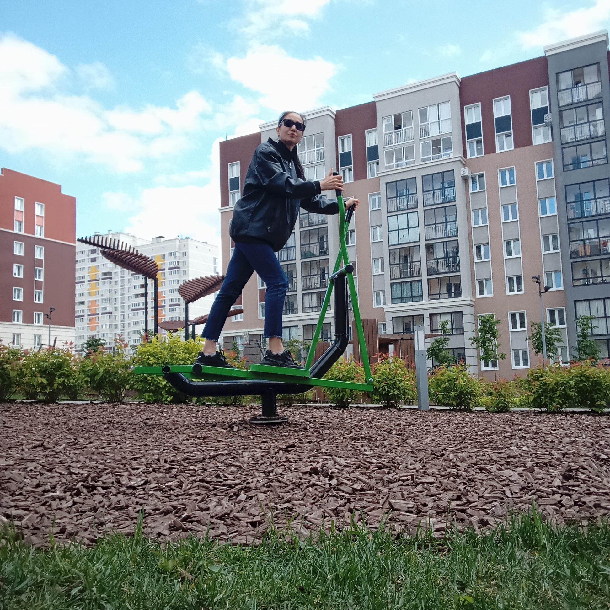 An outdoor eye-level shot captures a person exercising on a green and black elliptical trainer (air walker) in what appears to be a residential complex or public park in Pervomaysky, Russia. The person, an adult wearing sunglasses, a dark jacket, blue jeans, and black athletic shoes, is actively using the equipment, facing slightly left with a neutral expression.
The setting is bright and clear, under a blue sky with scattered white clouds, suggesting daytime. The foreground features a strip of vibrant green grass, transitioning to a wide area covered with dark brown bark mulch where the exercise equipment is located. Various green bushes and small trees are interspersed around the mulch area.
In the background, several modern, multi-story apartment buildings dominate the skyline. These buildings exhibit contemporary architecture with varied facades in shades of reddish-brown, gray, beige, and white, featuring numerous large windows and balconies. Another, more distant white building with colorful (orange, yellow) panels is visible on the left. Elements like wooden pergolas and pathway lights suggest a well-maintained outdoor communal space. No discernible text is visible in the image.