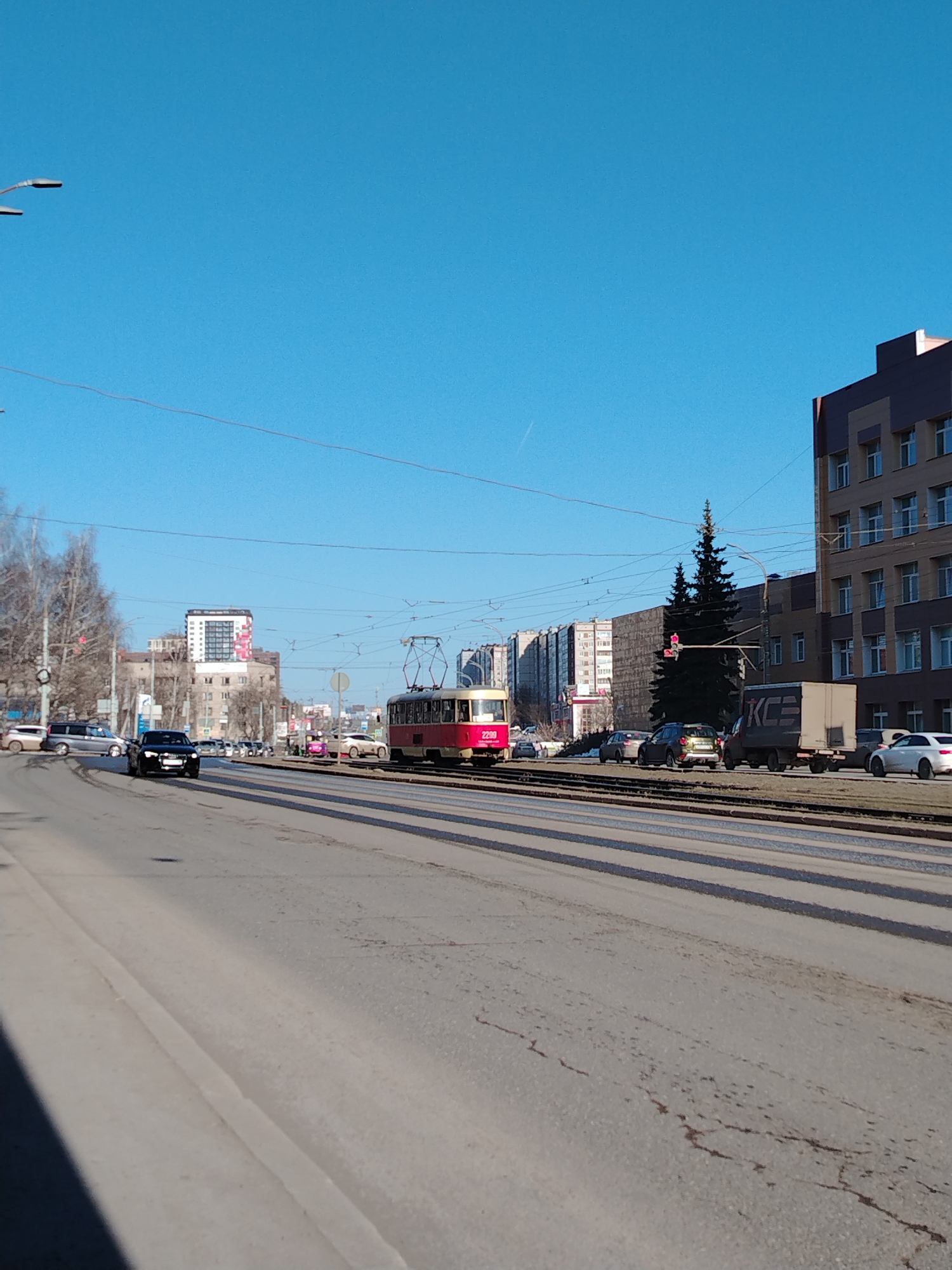 The image depicts a street scene in Izhevsk, Russia on a sunny day. A red and white tram is positioned in the center of the frame, traveling along tram tracks that run down the middle of a wide street. Several vehicles, including cars and a delivery truck, are present on the road alongside the tram. Buildings of varying heights are visible in the background. The sky is clear and blue. No people are clearly visible.