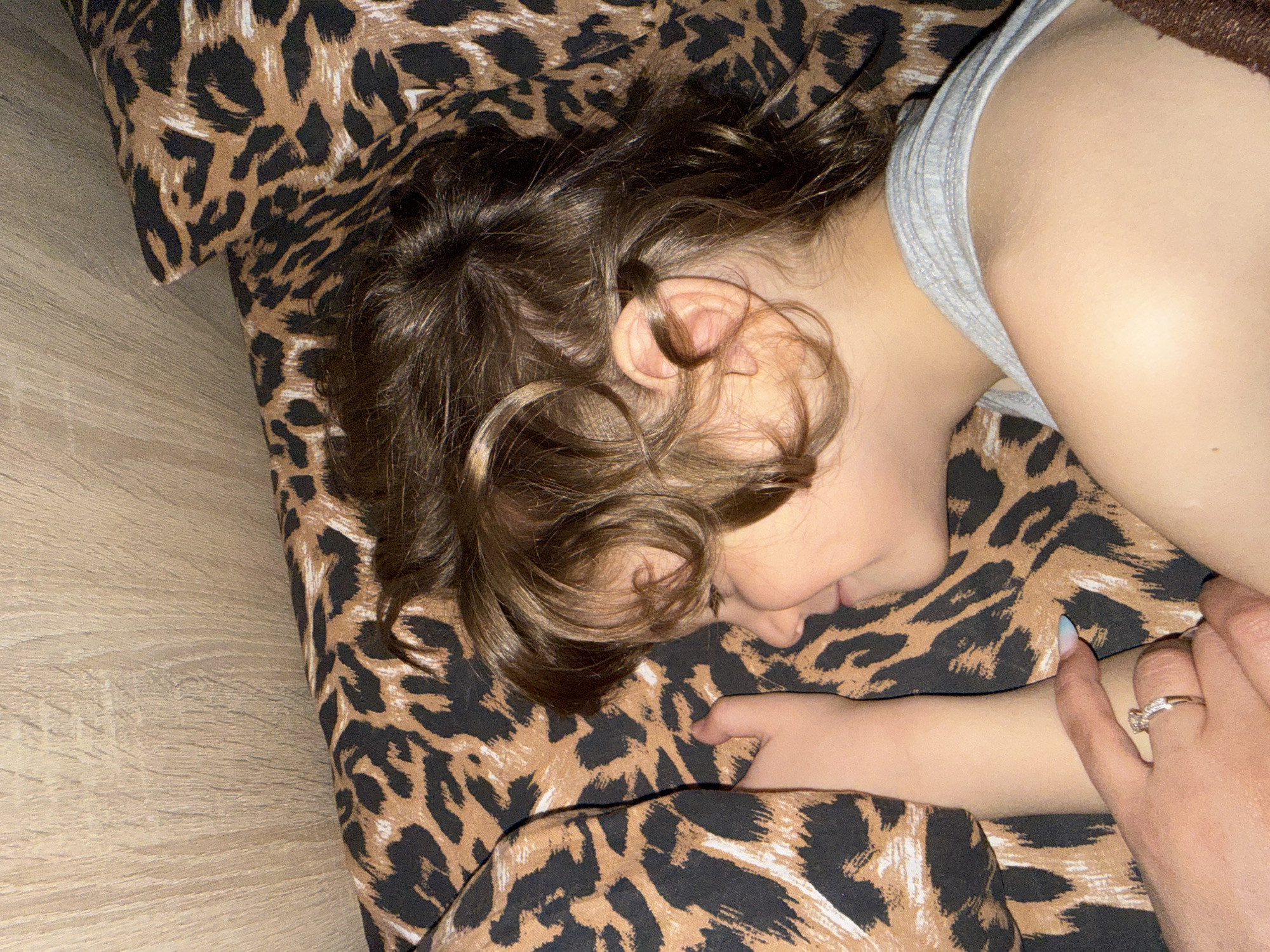 The image shows a close-up of a young person sleeping on a leopard-print surface, possibly a bed or sofa. The person is lying on their side with their head resting on a pillow that also has a leopard print. Their hair is brown and curly, and falls around their face. They are wearing a light grey tank top. One hand is visible, resting near their chest, adorned with a ring on one finger. The lighting suggests it might be nighttime or indoors. The overall mood is peaceful and relaxed, indicative of sleep. There is no visible text in the image.