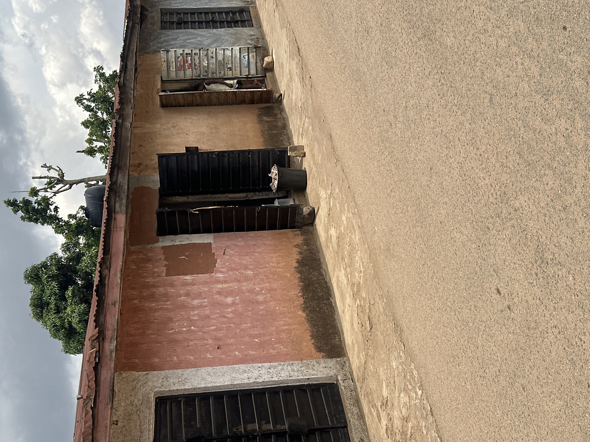 A street-level view shows a weathered, light-pink, single-story building with a corrugated metal roof. The building has several dark, slatted window or door openings. To the left, a large dark opening has horizontal slats. Next to it, a smaller opening shows a person, appearing to be a young woman or girl wearing a light-colored dress, standing in a doorway. Further to the right, another opening also has horizontal slats. A black trash can filled with debris is situated outside one of the openings. The ground in front of the building is a mix of dirt and cracked concrete, leading to an asphalt road. Trees and vegetation are visible behind the building, and the sky above is cloudy.
