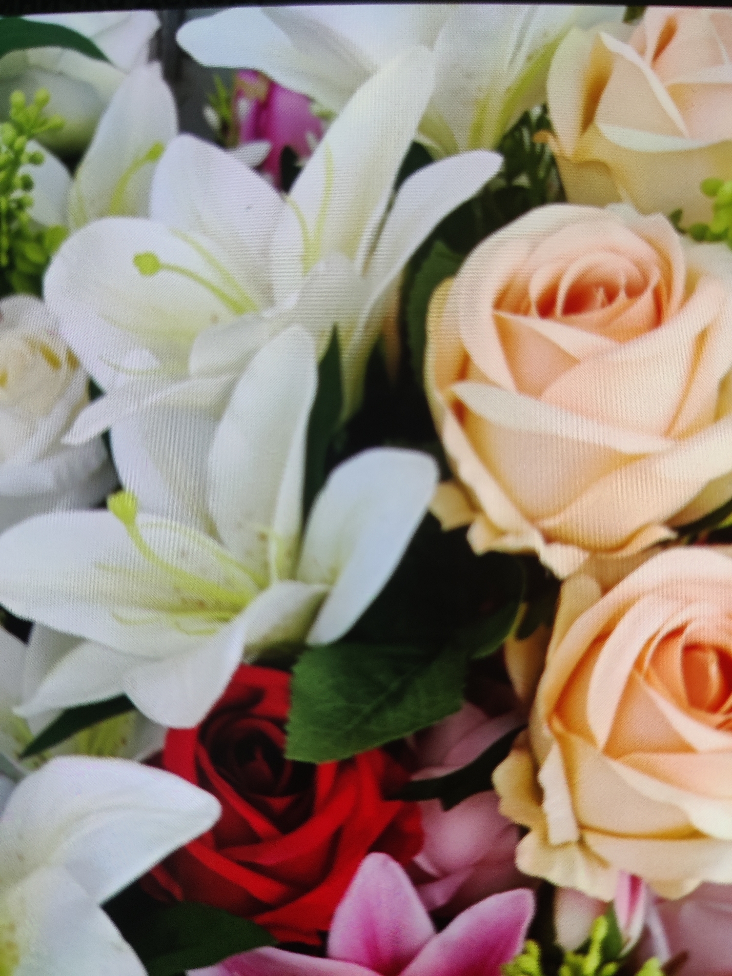 A close-up, overhead shot of a floral arrangement is featured. The bouquet is comprised of several types of flowers including peach roses, white lilies, and a single red rose. Green foliage and small buds are interspersed throughout the arrangement. The image is taken indoors, likely in a home or event setting. There are no people or actions depicted. The location of Lower Paxton Township, United States of America is not visually evident from the image.