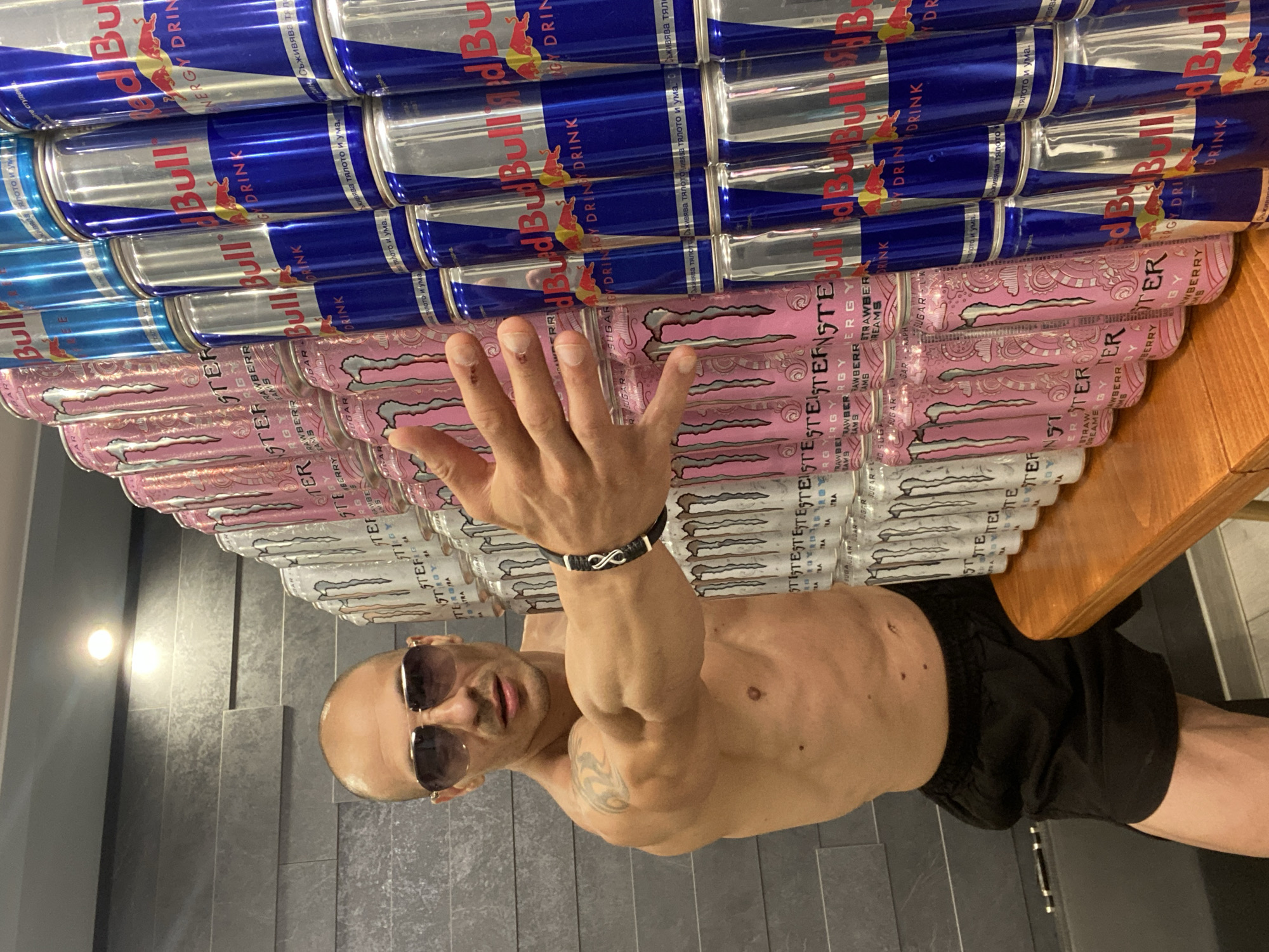 In Pleven, Bulgaria, a bald man with sunglasses and a muscular physique, shirtless and wearing black shorts, stands in front of a wall of stacked energy drink cans. The cans are a mix of pink Monster Energy drinks and blue Red Bull. The man's right arm is extended forward, touching the cans. The background appears to be an interior setting with a textured gray wall and a light fixture.