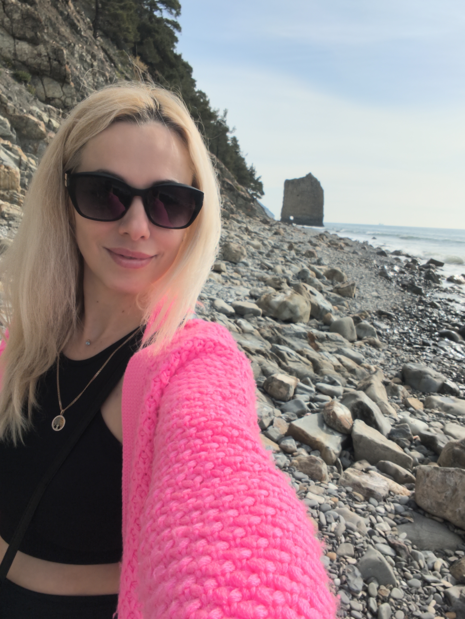 A woman with blonde hair, wearing dark sunglasses, a black top, and a bright pink sweater, is taking a selfie on a rocky beach. Behind her is the sea, a rock formation, and a rocky hillside with trees. This location is in Praskoveyevka, Russia.