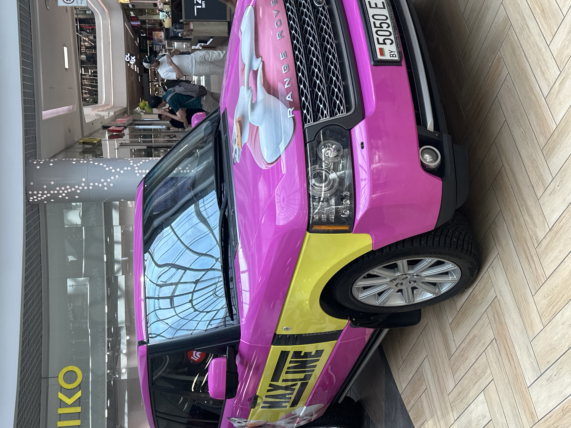 A bright pink Range Rover with yellow accents and a graphic of a duck wearing a pink shirt is parked on a herringbone-patterned wooden floor inside a modern shopping mall in Minsk, Belarus. The mall features a reflective ceiling with a grid pattern and a prominent "Seiko" sign. Several people are visible in the background, browsing shops and walking through the mall. The license plate of the Range Rover is visible and reads "5050 E BY".
