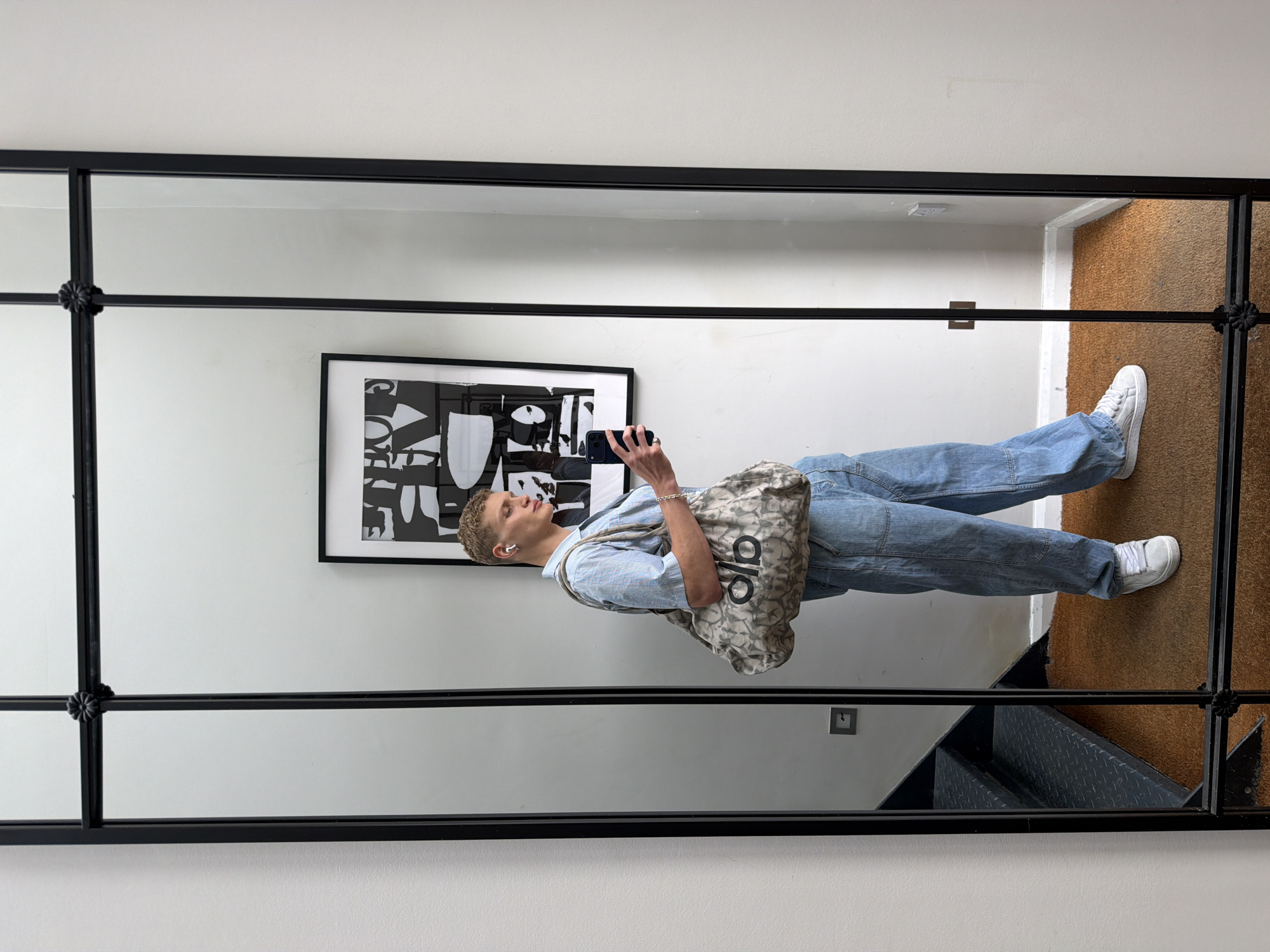 A person stands in front of a large, multi-paneled mirror, taking a selfie with a smartphone. They are wearing a light blue pinstriped short-sleeved button-up shirt, light blue baggy denim jeans, and white sneakers. A beige tote bag with a repeated logo pattern is slung over their left shoulder. They are also wearing white wireless earbuds. The background visible in the mirror includes a white wall with a framed black and white abstract art piece and a staircase with metal treads. The floor is covered with a brown mat. The overall scene suggests an indoor, residential setting, possibly in London, United Kingdom.