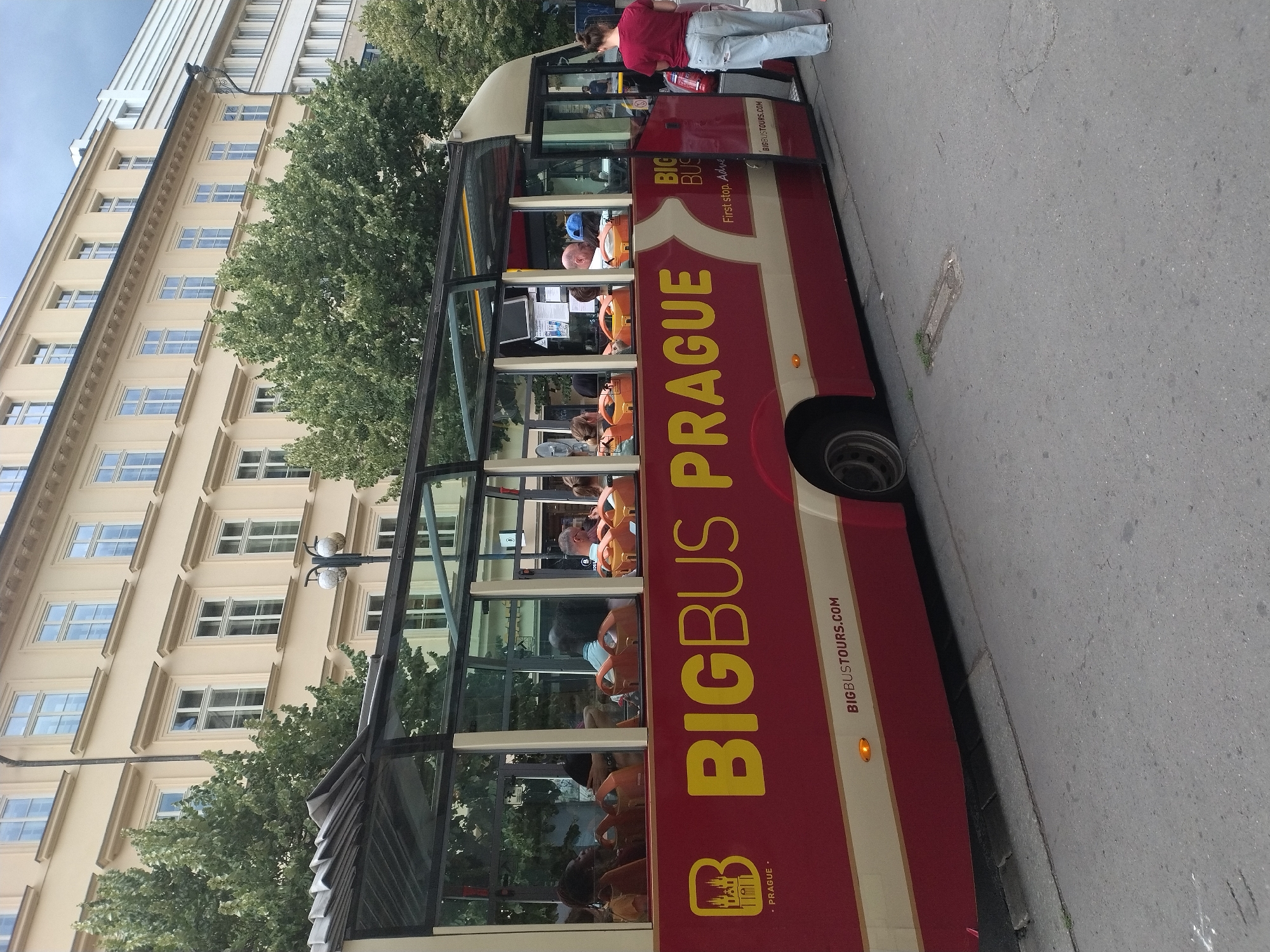 An open-top double-decker bus with the branding "BIGBUS PRAGUE" is parked on a cobblestone street in Prague, Czechia. The bus is red and cream with yellow lettering. Several passengers are seated inside the bus, some looking out the windows, while others are obscured by the angle. A woman in a red shirt and light blue jeans stands beside the open door of the bus, facing away from the camera. The bus is positioned in front of a cream-colored building with multiple windows and an ornate facade. Trees with green foliage are visible behind the bus and to the left. The sky is overcast.