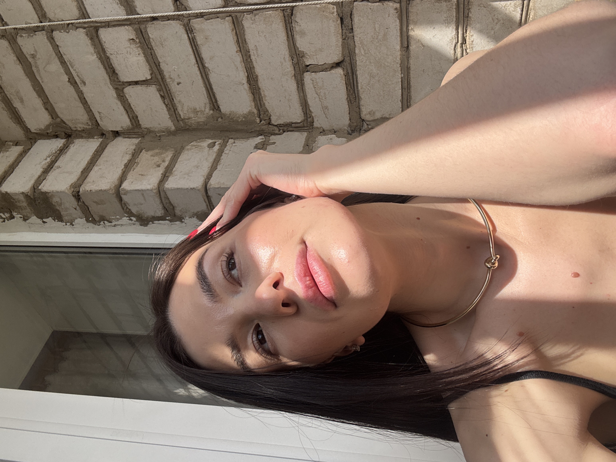 A woman with dark hair and fair skin is in an outdoor setting, possibly a balcony. The background shows a brick structure, likely part of a building. The woman is wearing a gold necklace and a dark top. The image is taken in the city of Chaikovsky, Russia. The woman's hand is near her head, and sunlight is illuminating her face.