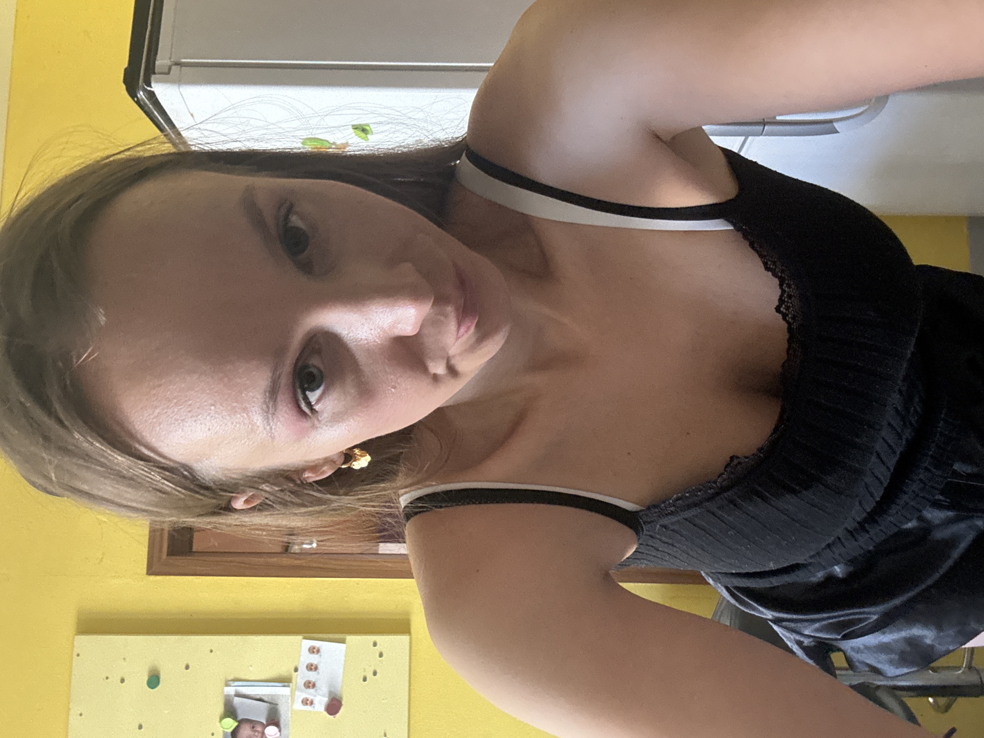 In a medium shot, a young woman with light brown hair pulled back is looking down and slightly to the left, her head tilted. She is wearing a black tank top with white trim and lace detailing along the neckline. A gold, flower-shaped earring is visible in her left ear. Her skin appears slightly shiny. To the left of the woman is a bright yellow bulletin board with several photos and colorful thumbtacks. Behind her, a wooden door and the side of a white refrigerator are visible. The overall scene suggests an indoor domestic setting.