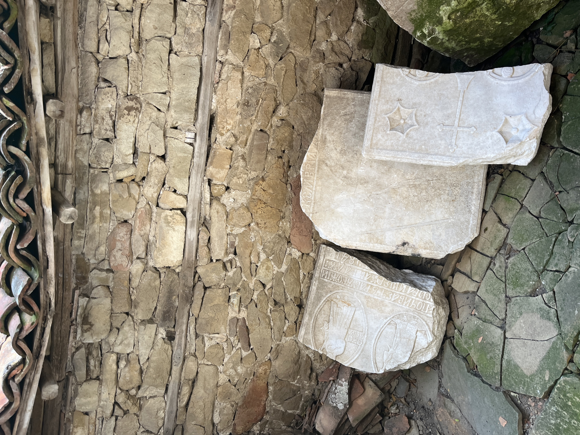 In Zheravna, Bulgaria, an outdoor scene depicts fragments of carved stone slabs leaning against a weathered, rough-hewn stone wall. The wall is constructed from irregular, tan and reddish-brown stones, with horizontal wooden beams reinforcing the structure. Above the wall, a section of a curved, tiled roof, possibly from an old building, is visible, with some tiles showing signs of age and moss growth.
In the foreground, several large, flat pieces of stone are arranged on a cracked, green-tiled surface that appears to be a floor or pavement. The stone fragments are intricately carved. One large slab, on the left, features circular engravings with figures and text in an ancient script. Another slab, to its right, is largely plain but has some faint text at the top. A smaller, upright slab on the right displays a cross and decorative star-like motifs. In the lower right corner, a large, moss-covered stone, possibly another fragment, is partially visible. The ground around the slabs is littered with smaller stone pieces and debris. The overall impression is of an archaeological site or a collection of historical artifacts stored outdoors.
