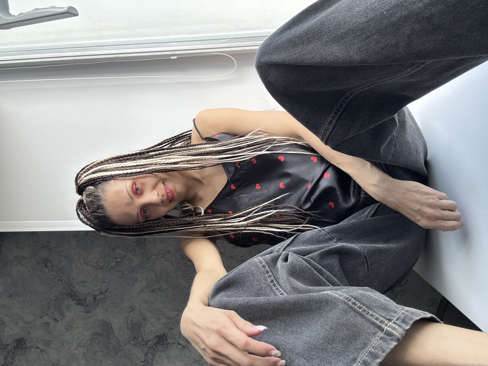 A woman is posing indoors, likely in a residential setting, in Rusanovo, Russia. She has long braids with blonde highlights and is wearing pink eyeshadow and red lipstick. She is wearing a black top with red hearts and baggy grey jeans. The background includes a white wall and a patterned grey surface. A window is visible at the top of the image.