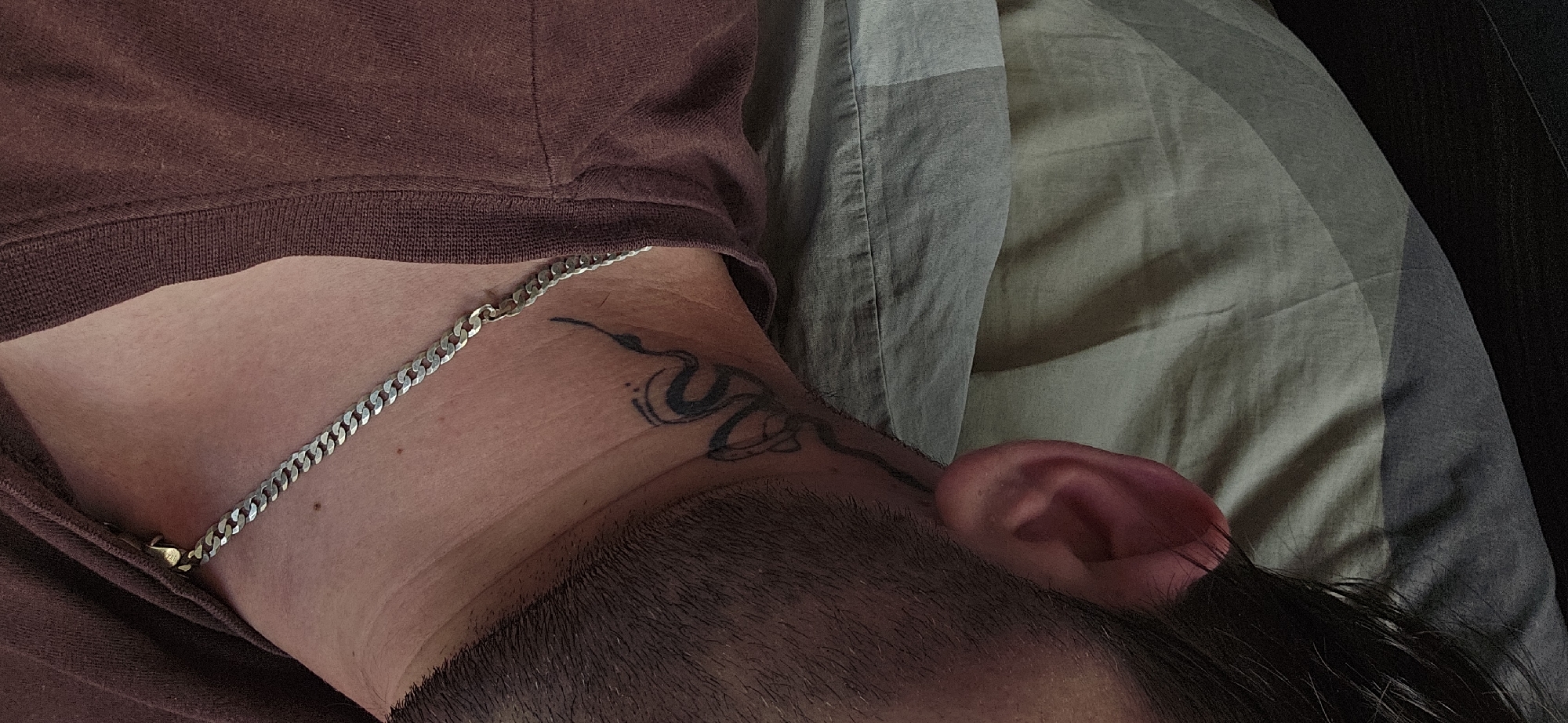 A close-up, aerial-perspective image shows a person lying on a bed with a grey and white patterned pillow visible on the right. The person wears a brown crewneck shirt and a silver chain necklace. A black tattoo is visible on their neck. The person's ear and a small portion of their face and hair are visible. The scene appears to be indoors. The location information (Kostenets, Bulgaria) does not seem relevant to the visible content.
