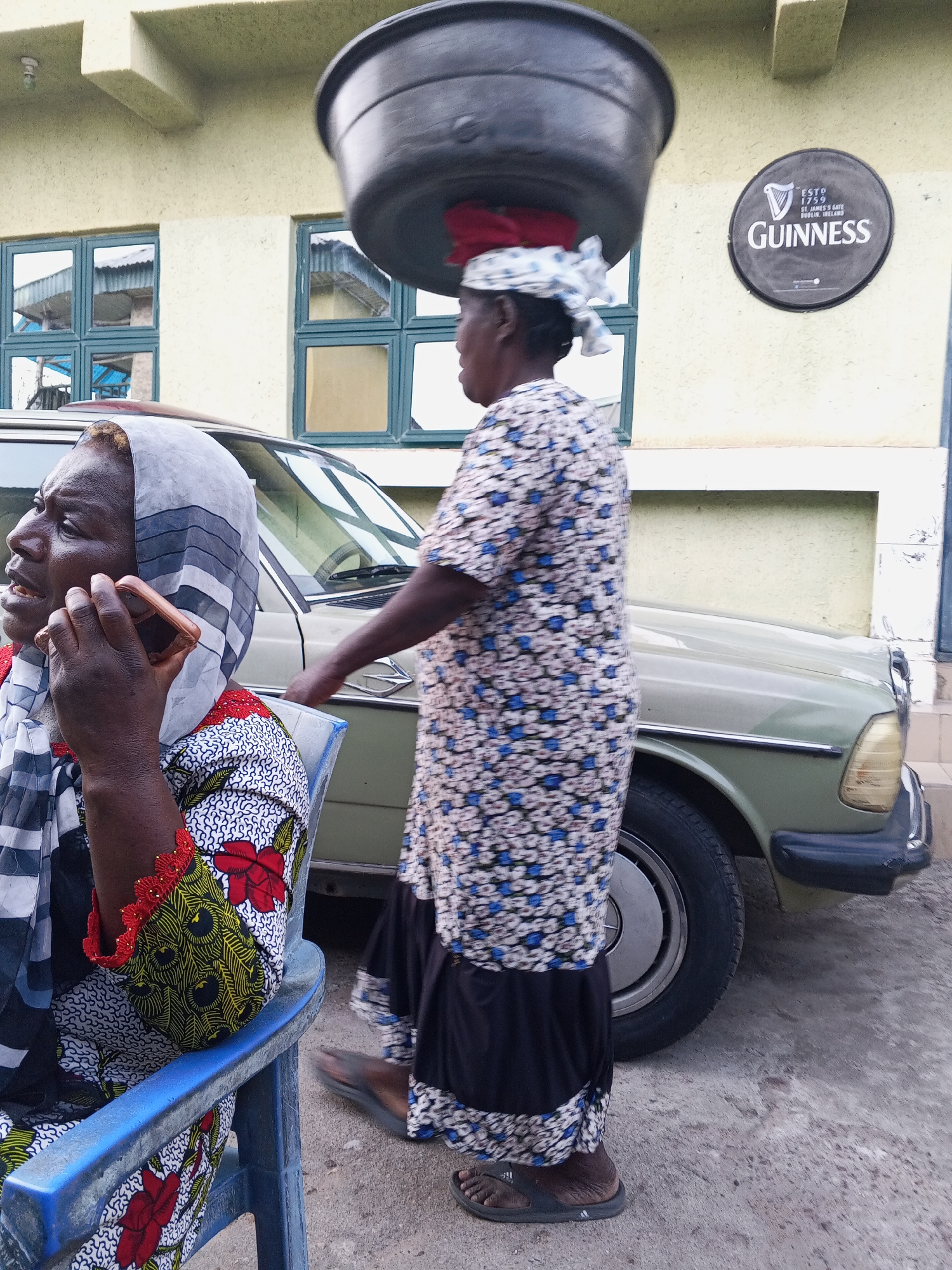 The image portrays a bustling street scene in Zango-ndaji, Nigeria, featuring two women and a vintage car against a building. In the foreground, an older woman with dark skin sits on a blue plastic chair. She is dressed in a vibrant, multi-patterned long-sleeved top adorned with red flowers and yellow/green motifs, paired with a black, white, and grey striped headscarf. She holds a gold-colored smartphone to her right ear, appearing deeply engrossed in a call.
Behind her, an older woman with dark skin walks from right to left, expertly balancing a large, dark basin on her head, cushioned by a red fabric. She wears a long-sleeved dress with a blue and white patterned top over a dark skirt and dark flip-flops, one bearing a visible "adidas" logo. Her profile is visible as she moves past.
A light olive-green or beige vintage sedan is parked between the two women and the building, serving as a backdrop. The building itself is light yellow or cream-colored, featuring multiple rectangular windows that reflect elements of the surroundings. A prominent black circular sign is mounted on the building wall, displaying the text: "ESTD 1759," "Guinness," and "St. James's Gate, Dublin, Ireland." The scene is brightly lit, indicating daytime, possibly under diffused light as harsh shadows are absent. The image captures a candid moment of daily activity and commerce in the Nigerian setting.