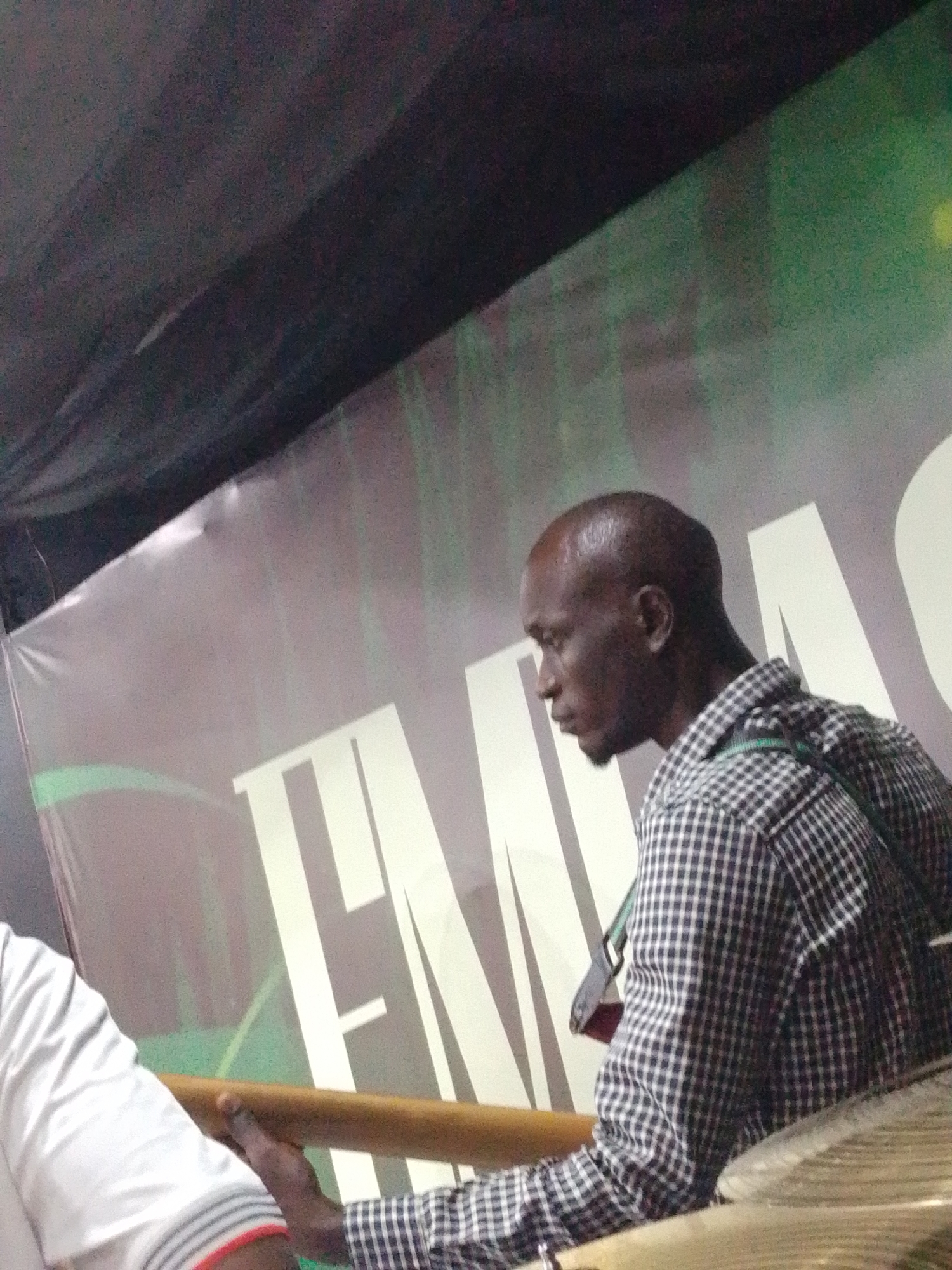 The image depicts a side profile of a bald Black man wearing a black and white checkered shirt and a green lanyard. He is seated, with his left arm resting on a wooden surface, possibly a table or railing. To his left, a portion of another person's arm is visible, wearing a white shirt with gray stripes and a red band around the wrist. The background features a large banner with bold white lettering that reads "EMAS", with a green pattern behind it. The scene appears to be indoors, possibly at an event or venue in Osogbo, Nigeria. The lighting suggests an artificial indoor environment.