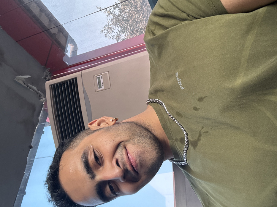 A man with short dark hair and a silver chain poses indoors with a slight smile. He wears an olive green t-shirt with a small logo, possibly “Pepe”, on the upper left. Behind him is a neutral-toned air conditioning unit with a digital display. Outside, through a window, a blurred view of a tree and the sky is visible. The image appears to be taken at an angle.