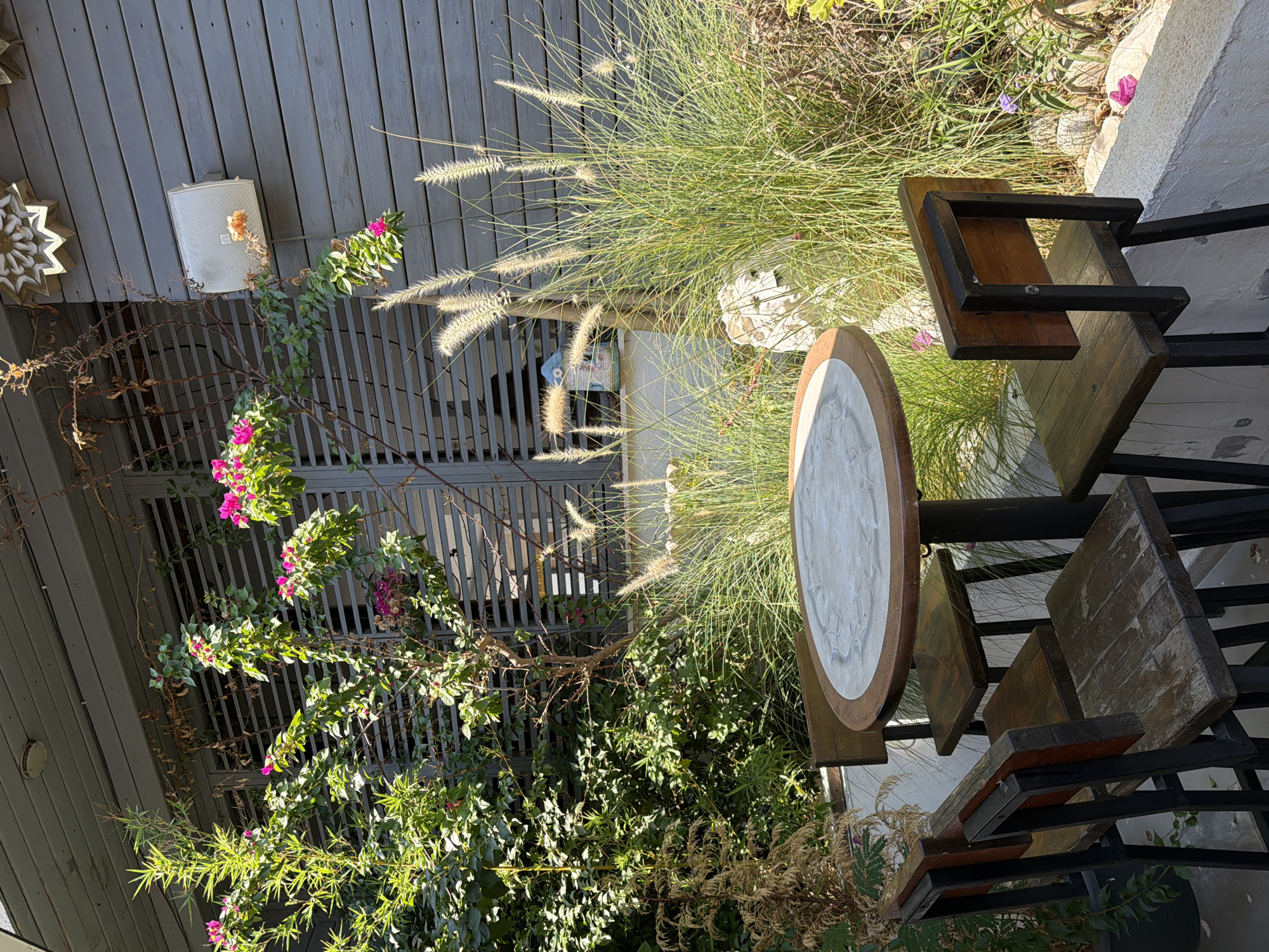 This image depicts an outdoor seating area with a round table and four wooden chairs. The table has a light-colored, possibly marble, top with a decorative pattern and a dark wooden rim. The chairs are made of dark wood and metal, with simple, rustic designs. They are arranged around the table.
The setting appears to be a garden or patio area, with lush green foliage and flowering plants in the background. Tall, feathery grasses are prominent, adding a touch of wildness to the scene. Behind the vegetation, a grey wooden wall is visible, featuring horizontal slats. On this wall, a white outdoor speaker is mounted, and a decorative, star-shaped object is affixed above it. A slatted window or screen is also visible behind the plants.
The overall impression is one of a peaceful, perhaps cafe or restaurant, outdoor space. The lighting suggests it is daytime, with sunlight casting shadows and highlighting textures. There are no people present in the image.
While the image itself does not provide specific geographical markers, if this scene were located in Marmaris, Turkey, it would likely be a charming spot within a coastal town known for its vibrant atmosphere and beautiful natural surroundings. Such a seating area could be found in a restaurant or cafe overlooking the Aegean Sea or nestled within the town's picturesque streets. The lush greenery suggests a Mediterranean climate, which is characteristic of Marmaris.
