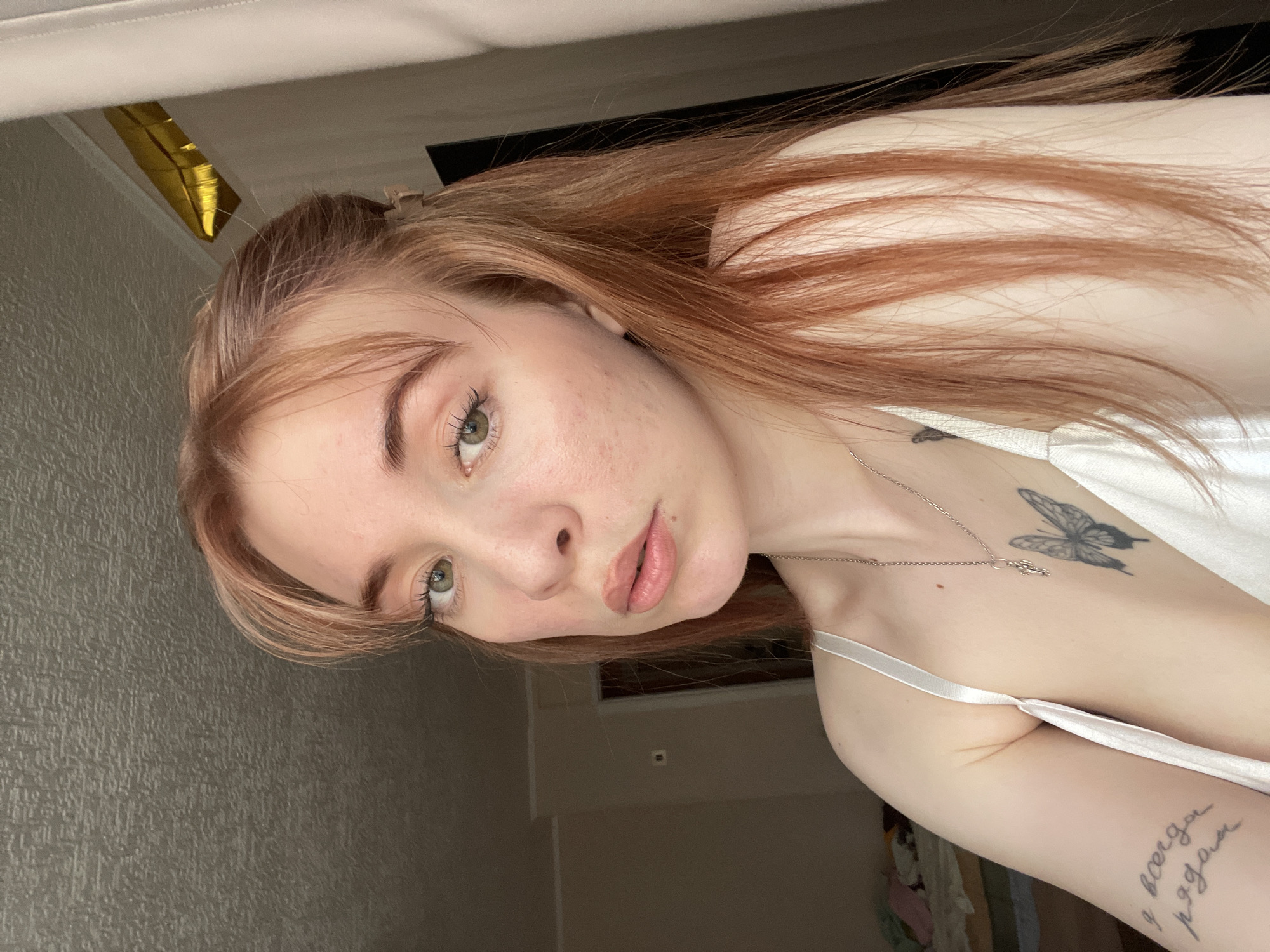 A close-up, slightly angled shot depicts a fair-skinned young woman with reddish-blonde hair. She has light eyes and a neutral expression. Part of a white garment with a thin strap is visible on her upper body. The woman has a tattoo of a butterfly on her upper chest and lettering on her upper arm. The background includes a textured wall and glimpses of a room with a balloon visible in the upper-left corner. The location is Unknown, Unknown.