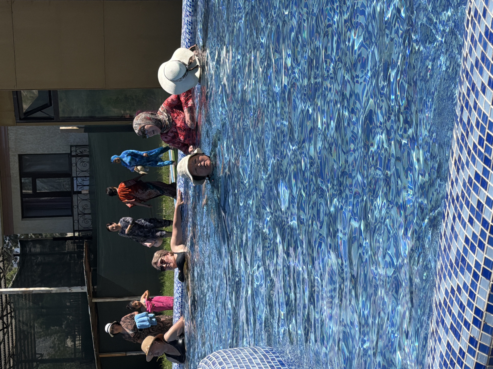 A group of individuals are gathered at the edge of a swimming pool. In the foreground, several people are partially submerged in the blue tiled pool. Two women are wearing swimsuits and hats, with one woman resting her arm on the pool's edge. Another woman, wearing a hijab and a red patterned top, is looking towards the left. A child with a hat is also in the pool. In the background, standing on the grass next to the pool, are several more individuals. Some are wearing traditional clothing, while others are in swim attire. The environment appears to be an outdoor setting with a building and some fencing visible behind the people. The water in the pool is rippling and reflects sunlight.