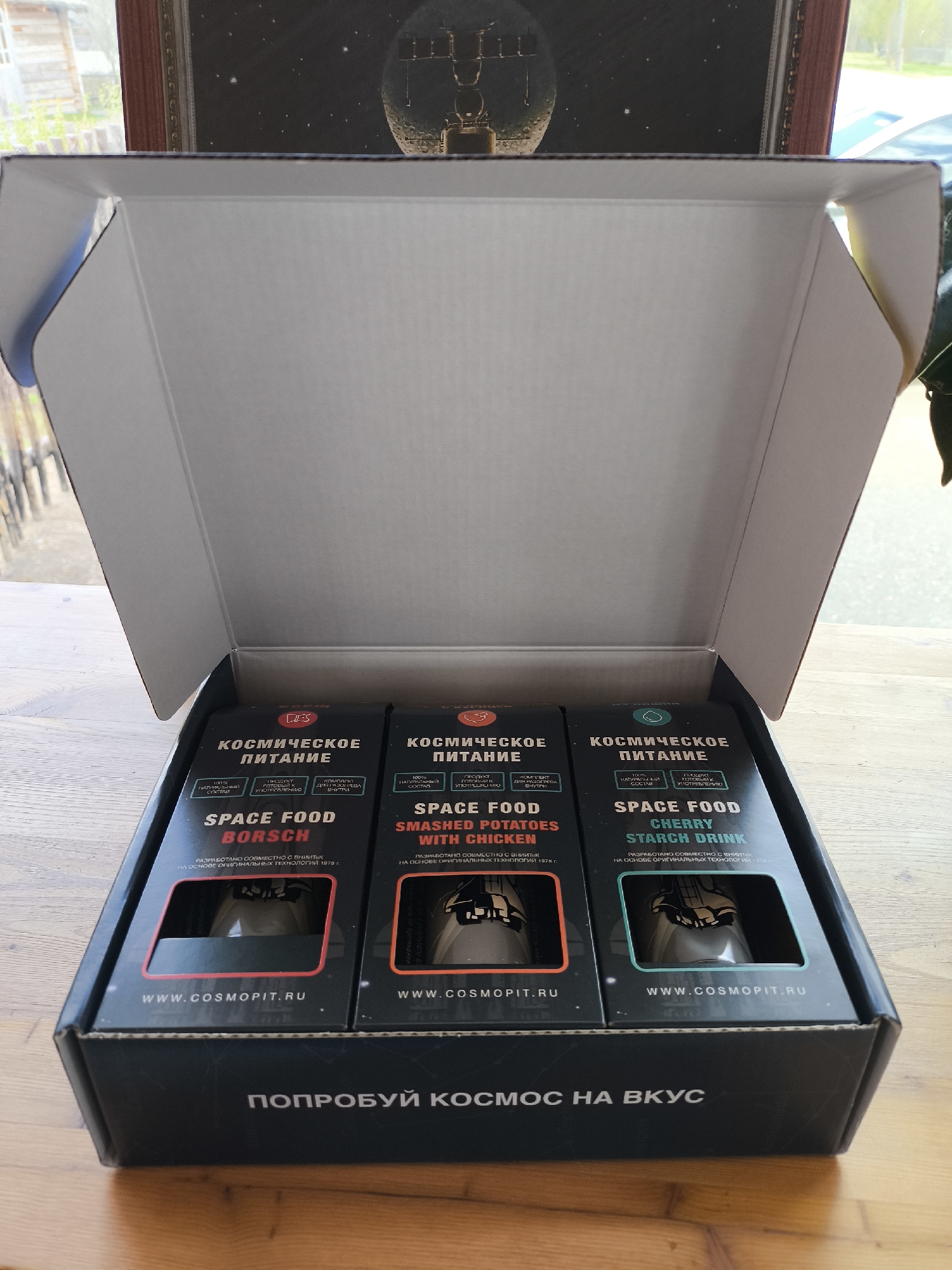 The image displays an open black cardboard box, resting on a light-colored wooden table, containing three novelty "Space Food" items. The interior of the box flaps is white. On the bottom exterior of the box, the Russian slogan "ПОПРОБУЙ КОСМОС НА ВКУС" (Taste Space) is visible.
Inside are three vertically arranged, dark-colored packages, each with a distinct color accent and a clear window revealing a tube-like container. From left to right, the items are: "SPACE FOOD BORSCH" (with a red accent), "SPACE FOOD SMASHED POTATOES WITH CHICKEN" (with an orange accent), and "SPACE FOOD CHERRY STARCH DRINK" (with a blue-green accent). Each package is prominently labeled "КОСМИЧЕСКОЕ ПИТАНИЕ" (Space Food) in Russian. Additional text on the packaging, both in Russian and English, highlights features such as "100% натуральный состав" (100% natural composition), "готовый к употреблению" (ready to eat), and states that the products were "РАЗРАБОТАНО СОВМЕСТНО С ВНИИТЕК НА ОСНОВЕ ОРИГИНАЛЬНЫХ ТЕХНОЛОГИЙ 1978 г." (Developed jointly with VNIItek based on original technologies of 1978). All packages also feature the website "WWW.COSMOPIT.RU".
The background shows a blurred outdoor scene through a window, featuring green foliage and distant structures, indicating daytime. Above the box, a dark-framed artwork depicting a celestial body with structures against a starry sky reinforces the space theme. The scene suggests an indoor presentation of these space-themed food products, likely in Kluschino, Russia, given the language on the packaging.