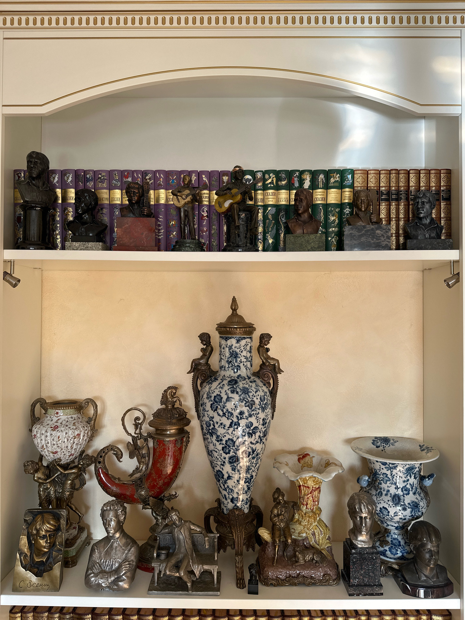 This image depicts a close-up, eye-level view of a white, built-in shelf unit, adorned with gold trim along the top. The shelf unit is divided into two tiers.
The upper tier displays a collection of decorative items and books. On the left, there are several bronze busts of men, each mounted on a pedestal. Behind the busts, a row of books with purple and green spines, some featuring gold lettering and ornate designs, lines the shelf. Scattered amongst the books and busts are further bronze figurines of men playing musical instruments. The right side of the upper shelf holds more bronze busts and a selection of books with patterned gold and brown spines.
The lower tier of the shelf unit is a backdrop of textured, light beige wallpaper. In the foreground of this lower tier, a collection of varied decorative objects is arranged. On the far left is an ornate, multi-handled vase with a floral pattern. To its right sits a red, horn-shaped decorative piece with intricate metalwork and figures. Center stage is a large, tall vase with a blue and white floral pattern. To the right of this vase are two smaller blue and white patterned vases, one with a wide opening. Between these vases, and to the left of the smaller vases, are several bronze figurines depicting human figures, some interacting with animals or objects.
The image does not contain any information to suggest its location in Murmansk, Russia.