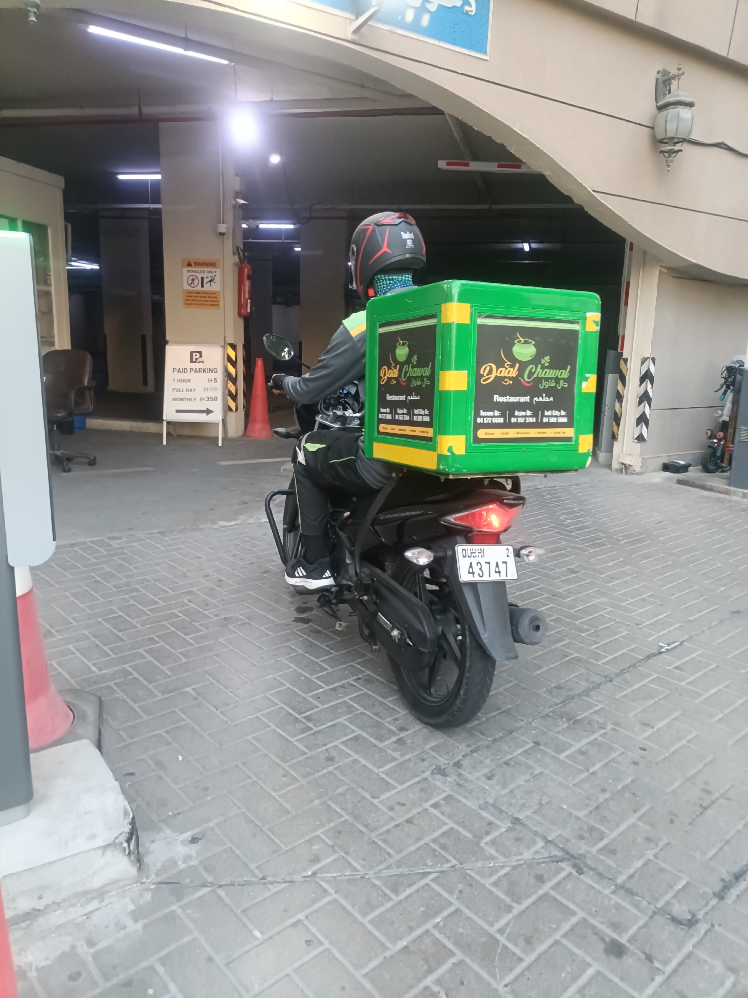 In Dubai, United Arab Emirates, a delivery person on a motorcycle navigates a paved area, possibly a parking lot or driveway, within a covered structure. The motorcycle, a black model, is fitted with a large, bright green delivery box on the rear, emblazoned with the logo "Daal Chawal Restaurant" in both English and Arabic. The delivery person is wearing a black and grey riding suit and a black helmet with red accents. In the background, a parking sign indicates rates for hourly, full-day, and monthly parking, along with a warning sign for vehicles. Other elements in the environment include concrete pillars, lighting fixtures, and a red traffic cone. The overall scene depicts a typical urban commercial delivery operation.