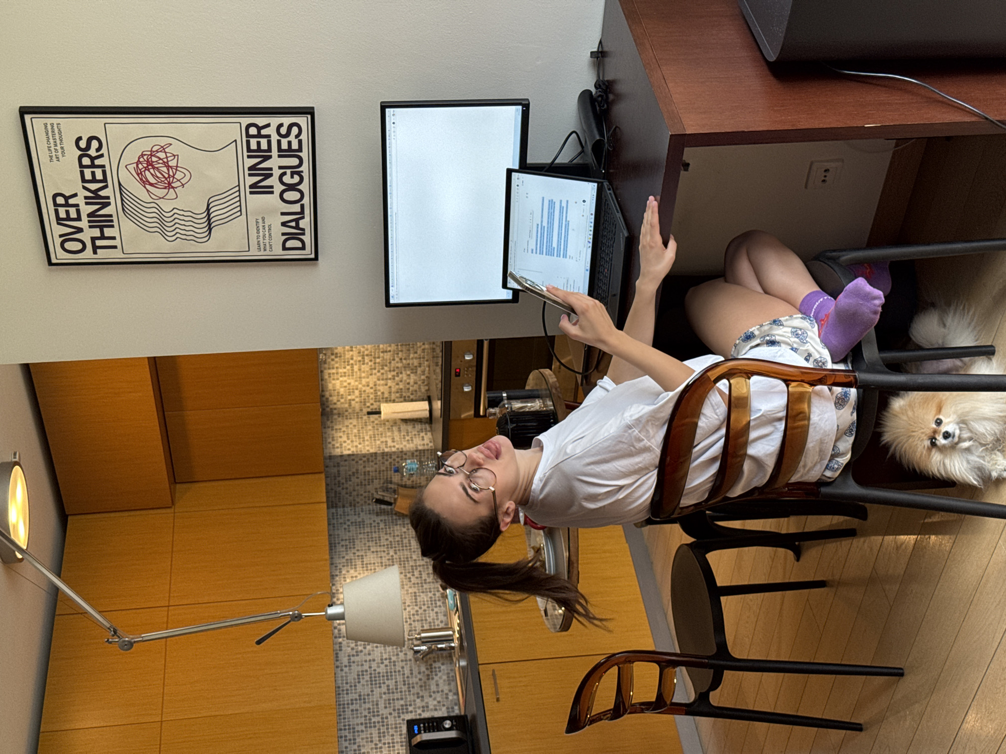 A woman sits in a brown chair in front of a desk with a computer and monitor. She is wearing glasses, a white shirt, and flowered shorts with purple socks. She is holding a cell phone in her left hand and raising her right hand. Beside her feet is a small, light-colored dog. Behind her is a kitchen with light wood cabinets and tile backsplash. On the wall to the left is a modern lamp, and to the left of the desk is a framed poster with the words "OVER THINKERS" and "INNER DIALOGUES" printed on it. The city of Unknown, Unknown is not specified in this image.