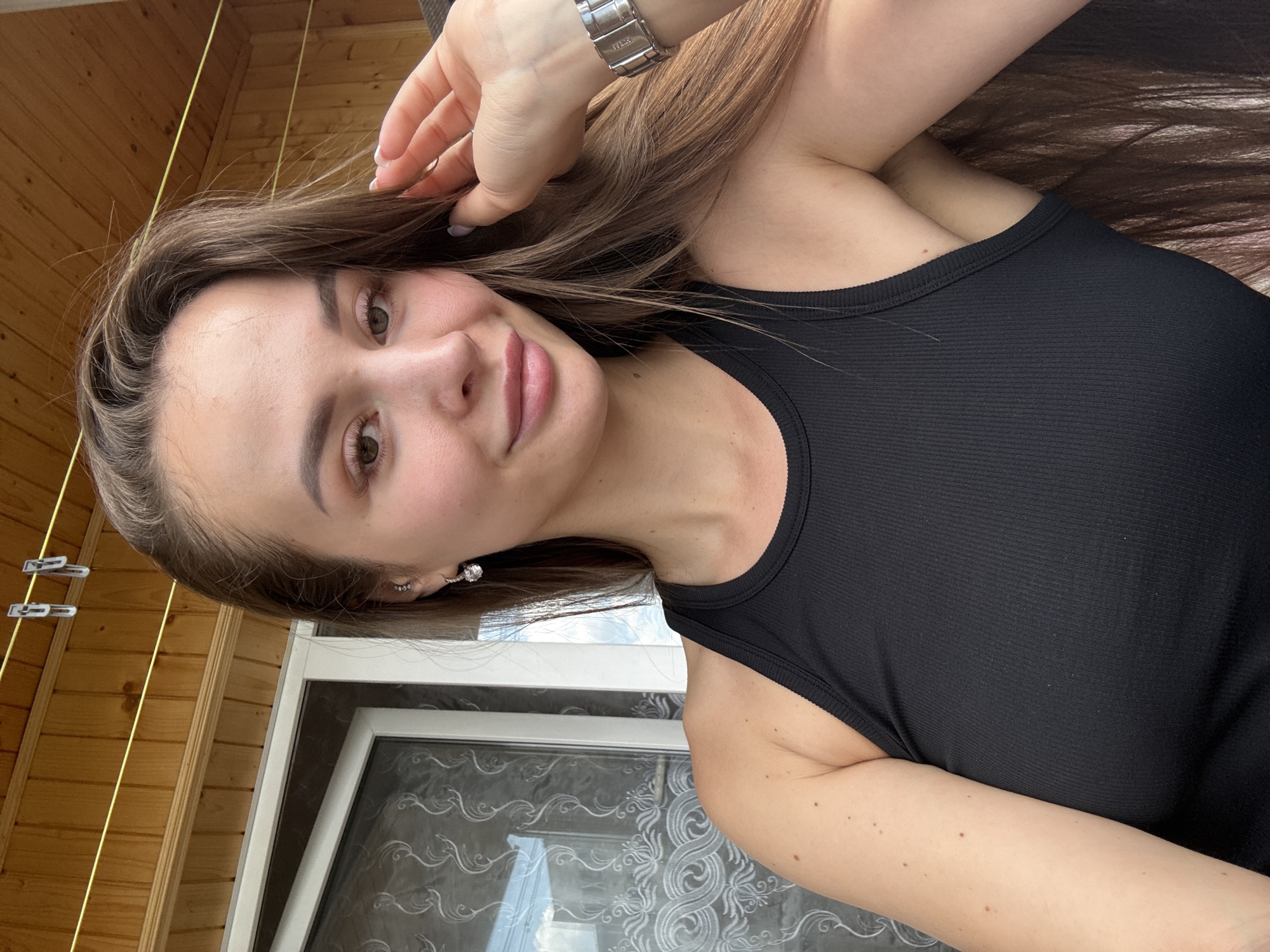 The image is a selfie of a young woman with long brown hair, wearing a black tank top and silver earrings. She is outside, with a wooden ceiling and part of a window visible in the background. There are clotheslines strung across the wooden ceiling. The lighting suggests it is daytime. There is no visual information in the image that indicates it is located in Kyiv, Ukraine.