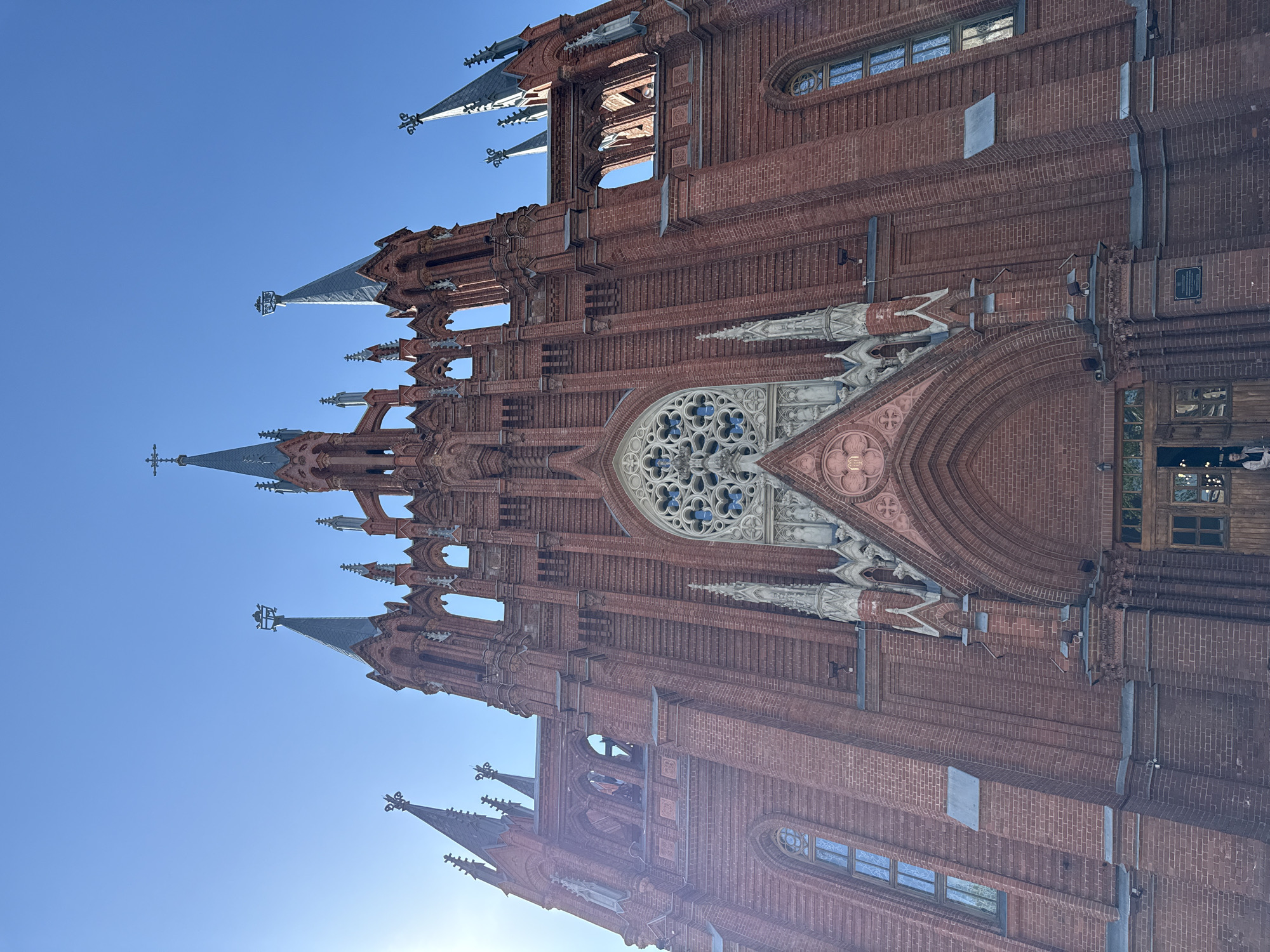 The image shows the Catholic Cathedral of the Immaculate Conception in Moscow, Russia, angled upwards against a clear blue sky. The cathedral is constructed of red brick and features ornate Gothic Revival architectural details, including spires, pointed arches, and a large rose window. There are no people visible in the main view of the cathedral, but two people are barely visible entering the building on the far right of the image.
