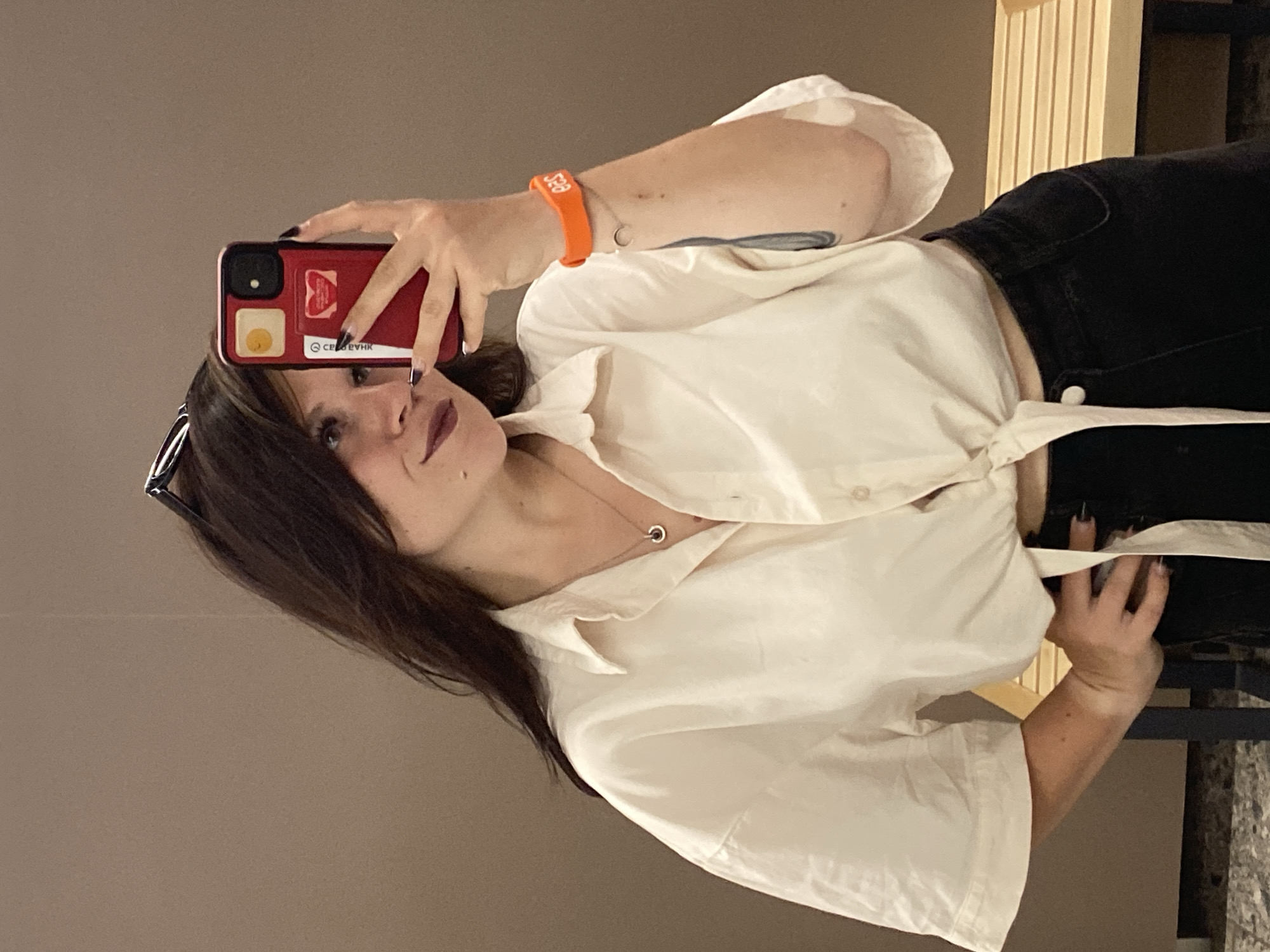 A young woman with long, dark hair and dark lipstick takes a selfie in what appears to be a fitting room. She is wearing a cream-colored, tied-front button-down shirt and dark pants. A red phone is held in her right hand, and an orange bracelet is on her left wrist. A silver necklace with a circular pendant is visible around her neck. The background is a plain, neutral wall. The image does not provide any specific details to indicate a location in Votkinsk, Russia.