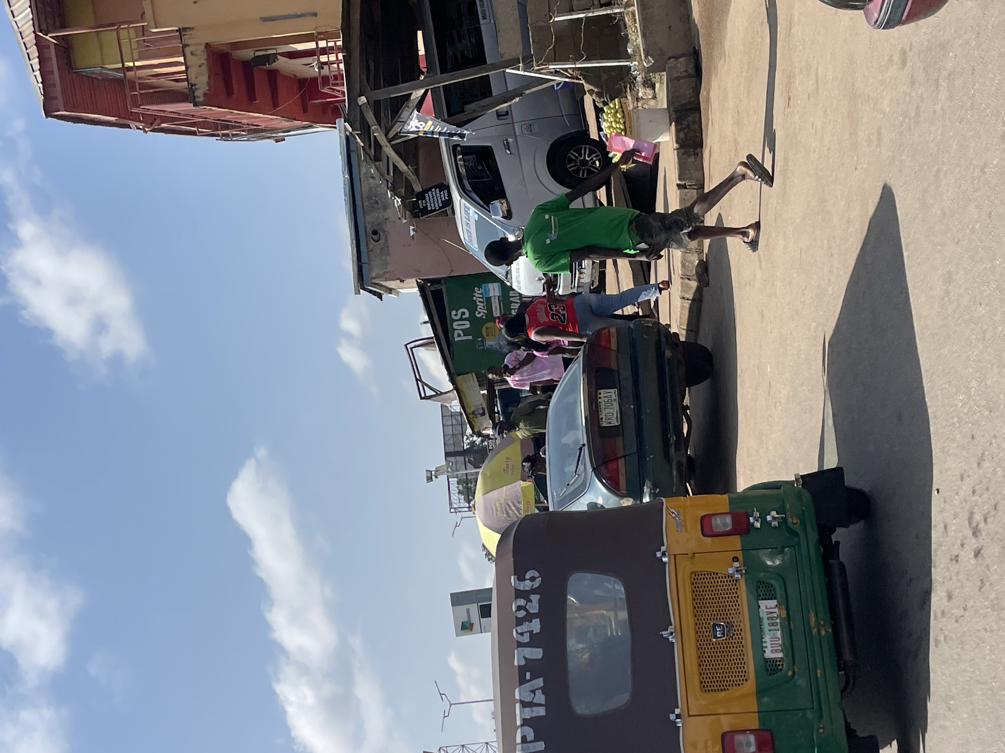 An outdoor, street-level, medium-shot photograph captures a bustling scene in Jos, Nigeria, on a bright, sunny day. The perspective is from behind a yellow and green autogyro in the foreground, which has the license plate "RUJ-188VE" and "PTA-7426" visible.
Several individuals are present. A man wearing a green t-shirt and camouflage shorts walks away from the viewer towards the right, carrying a pink object and a white bucket filled with fruit. Behind him, a silver van with the words "POWER IS LIFE" on its side is parked. Further back, a dark green sedan with the license plate "KRO-706AV" is also parked, and a woman in a pink shirt and jeans stands near it. A yellow and purple umbrella is visible behind the sedan.
In the background, various structures line the street. A multi-story building with red and yellow exterior paint and exposed stairs is on the right. To the left of that, a green building with "POS Sprite" signage and another sign with "BRAND" visible can be seen. Other makeshift structures and overhead wires contribute to the urban environment. The sky is blue with scattered white clouds. The overall atmosphere suggests a lively marketplace or commercial area.