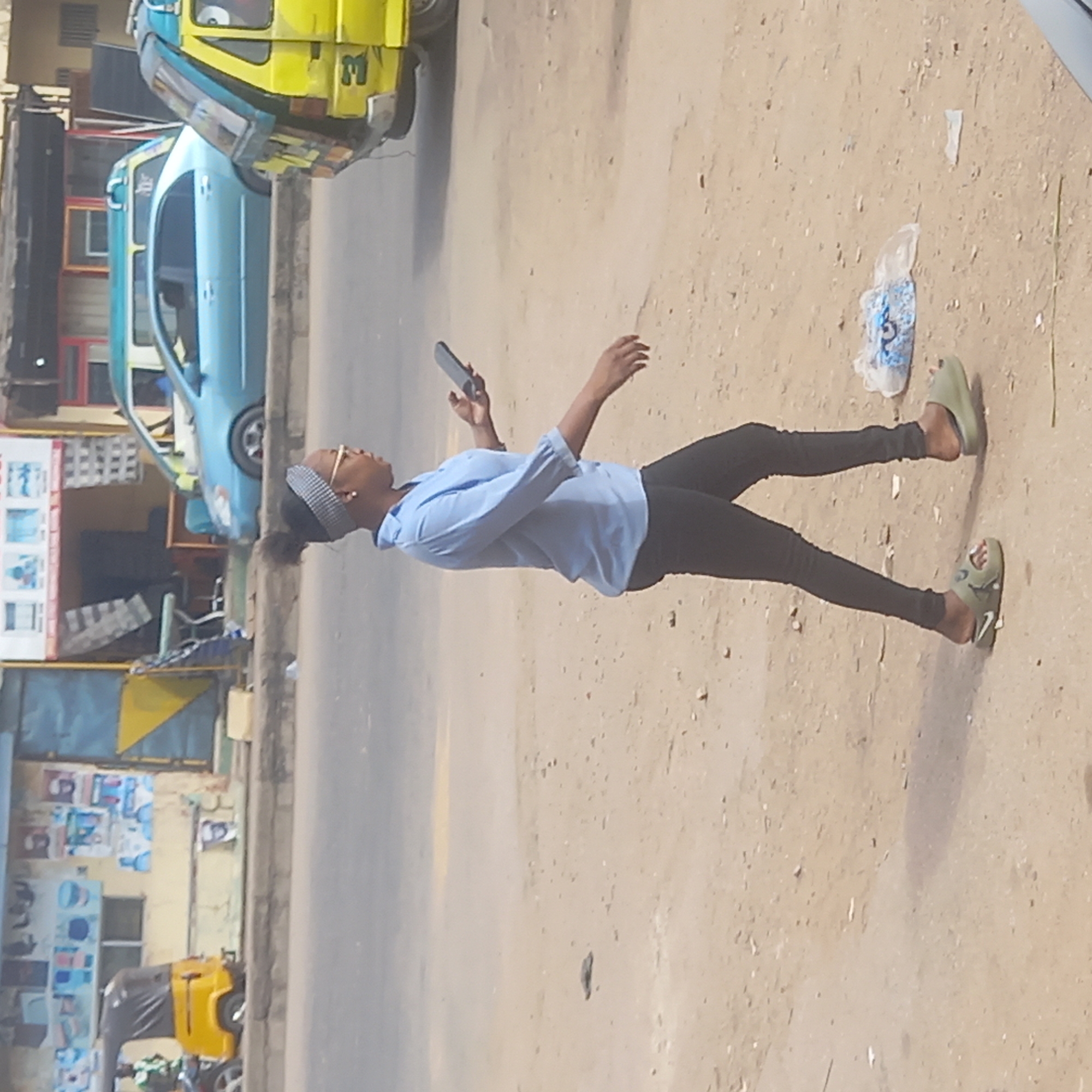 A young woman stands on a dirt-covered street in Dadi Riba, Nigeria. She is wearing a light blue shirt, black pants, and green sandals, and has a gray headband. She is holding a cell phone in her left hand and appears to be looking at it. The environment shows a street with parked or moving vehicles, including a yellow and blue three-wheeled vehicle. There is litter, including a plastic bag, on the ground near her feet. Buildings can be seen in the background with posters and advertisements displayed on the walls.