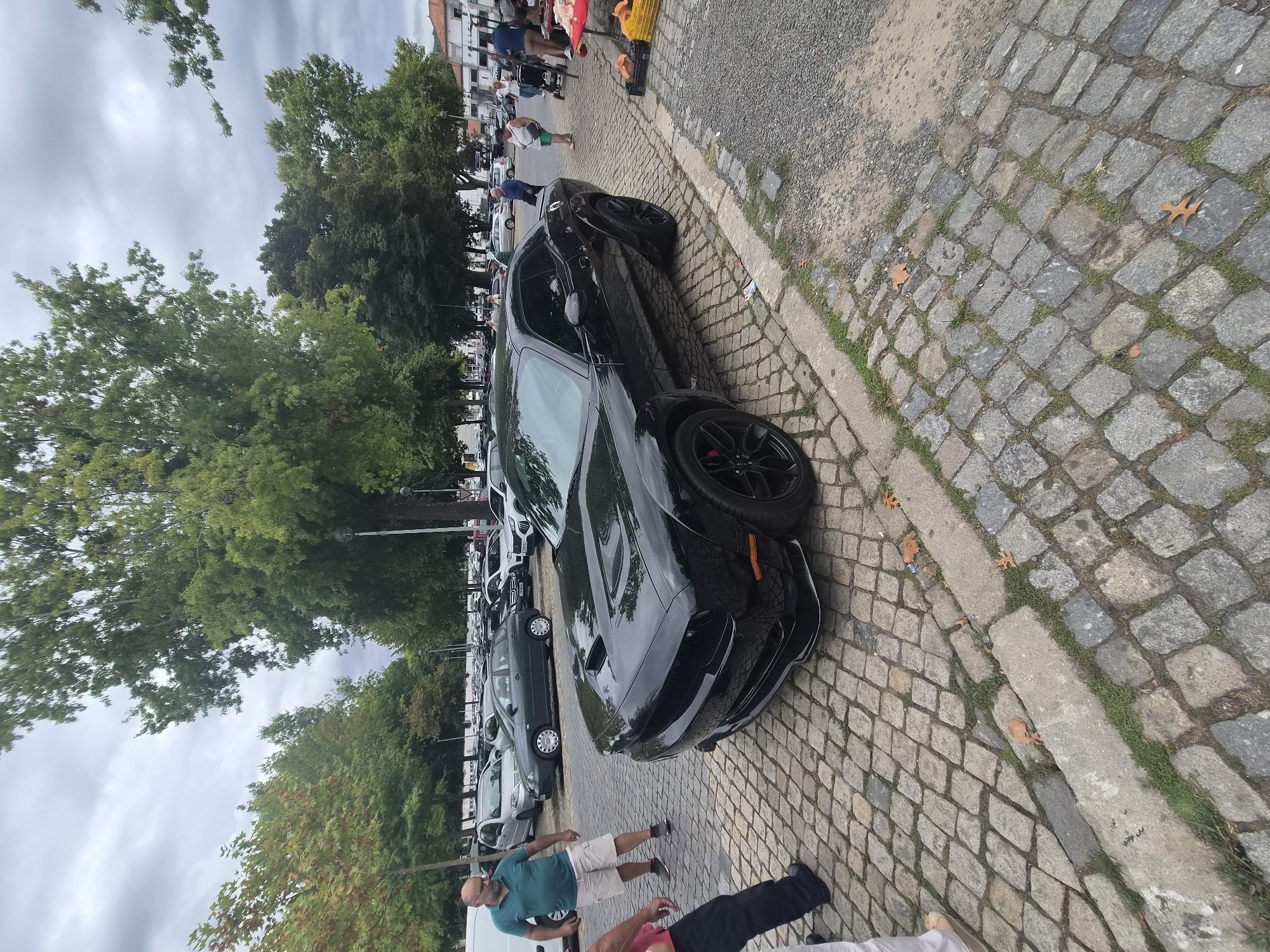 A black Dodge Challenger is parked on a cobblestone street in Barcelos, Portugal. The car is angled with its front passenger side facing the viewer. The street is lined with trees, and several people are visible in the background, some standing near other cars and a market stall. The sky is overcast. The car has a hood scoop and black rims. A man in a teal polo shirt and khaki shorts is standing to the left of the car, looking towards it.