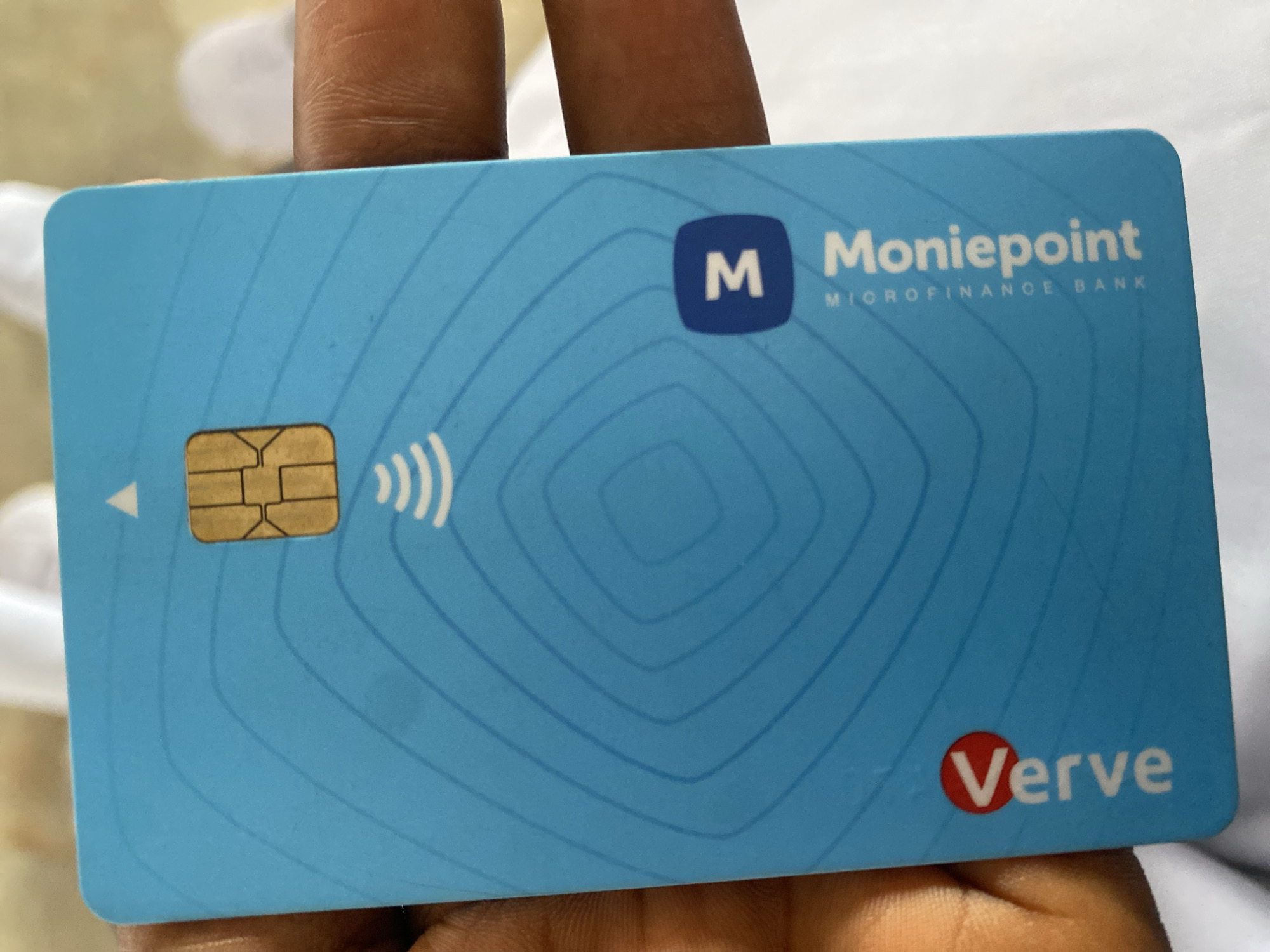 A close-up, high-angle shot shows a blue Moniepoint Verve debit card held by a person's fingers. The card features a gold chip at the top, a contactless payment symbol below it, and a pattern of concentric blue lines. The Moniepoint logo, an 'M' inside a blue square, is visible on the right side of the card, with the text "MONIEPOINT MICROFINANCE BANK" below it. The Verve logo, a red circle with white text, is on the bottom left. The background is out of focus, showing hints of white and skin tones. The image was taken in Jimeta, Nigeria.