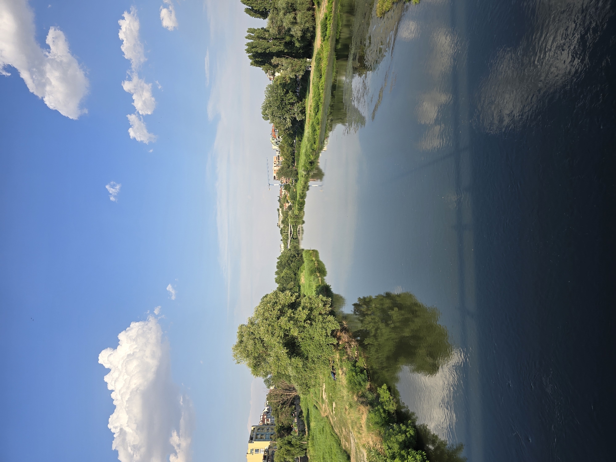 A vertical, eye-level, wide-angle shot captures a tranquil river flowing through a city, with buildings and lush greenery lining its banks. The sky is a brilliant blue, dotted with fluffy white clouds. The calm water mirrors the sky and the surrounding trees, creating a serene and picturesque scene. In the distance, a bridge spans the river, and construction cranes hint at ongoing development. The overall impression is one of a peaceful urban landscape with a harmonious blend of nature and civilization.