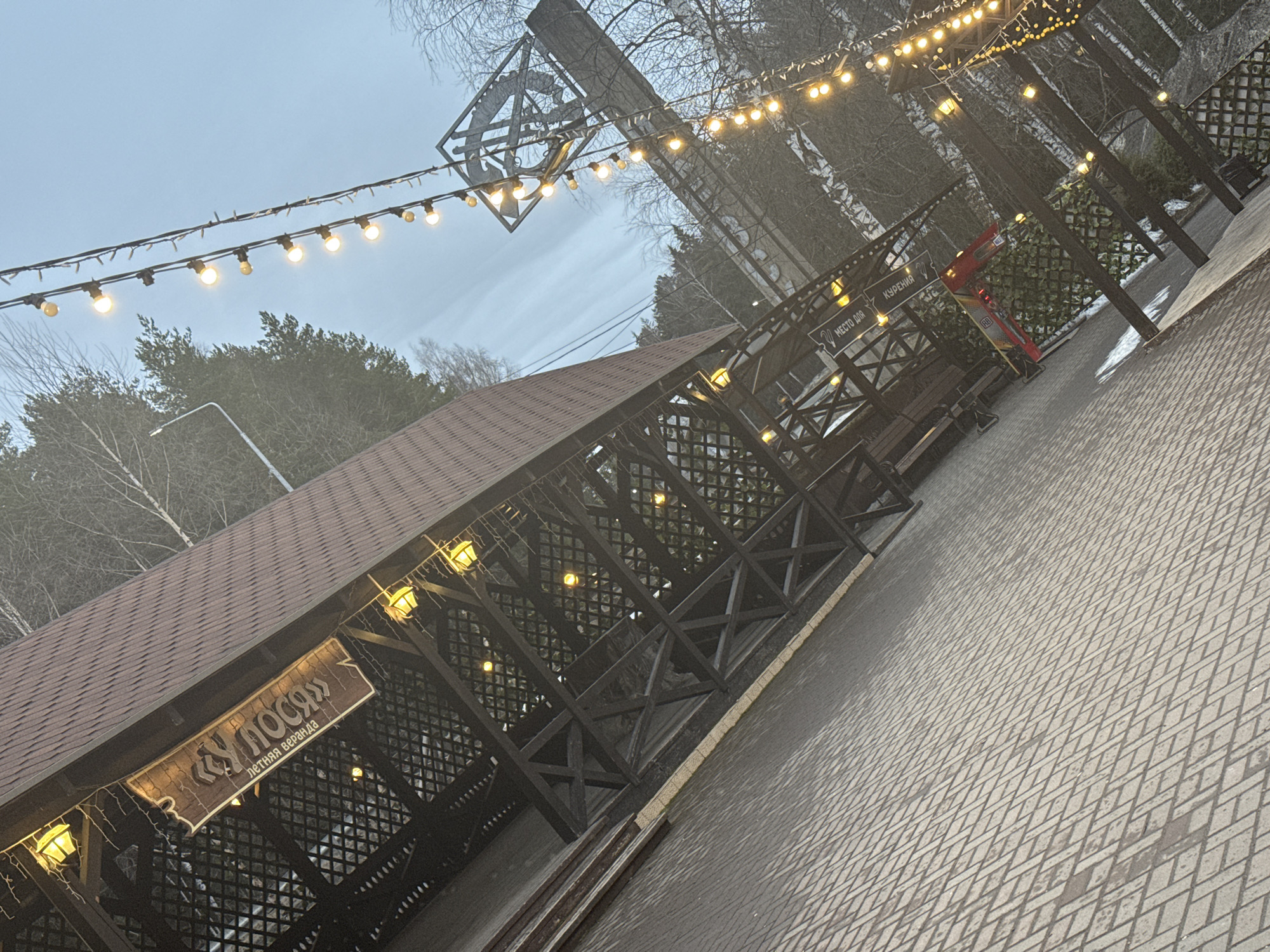 The image shows a partially covered outdoor terrace and patio area in Izhevsk, Russia. The terrace features a wooden structure with a brown tiled roof and decorative lattice panels. A sign with Cyrillic text identifies the venue as "У ЛОСЯ" (U Losya) and indicates it's a summer veranda. Strings of lights are strung across the terrace and overhead, adding illumination. Adjacent to the terrace is a paved patio area with a diamond pattern. Trees are visible in the background, suggesting a park or wooded area. The overall scene appears to be at dusk or early evening.