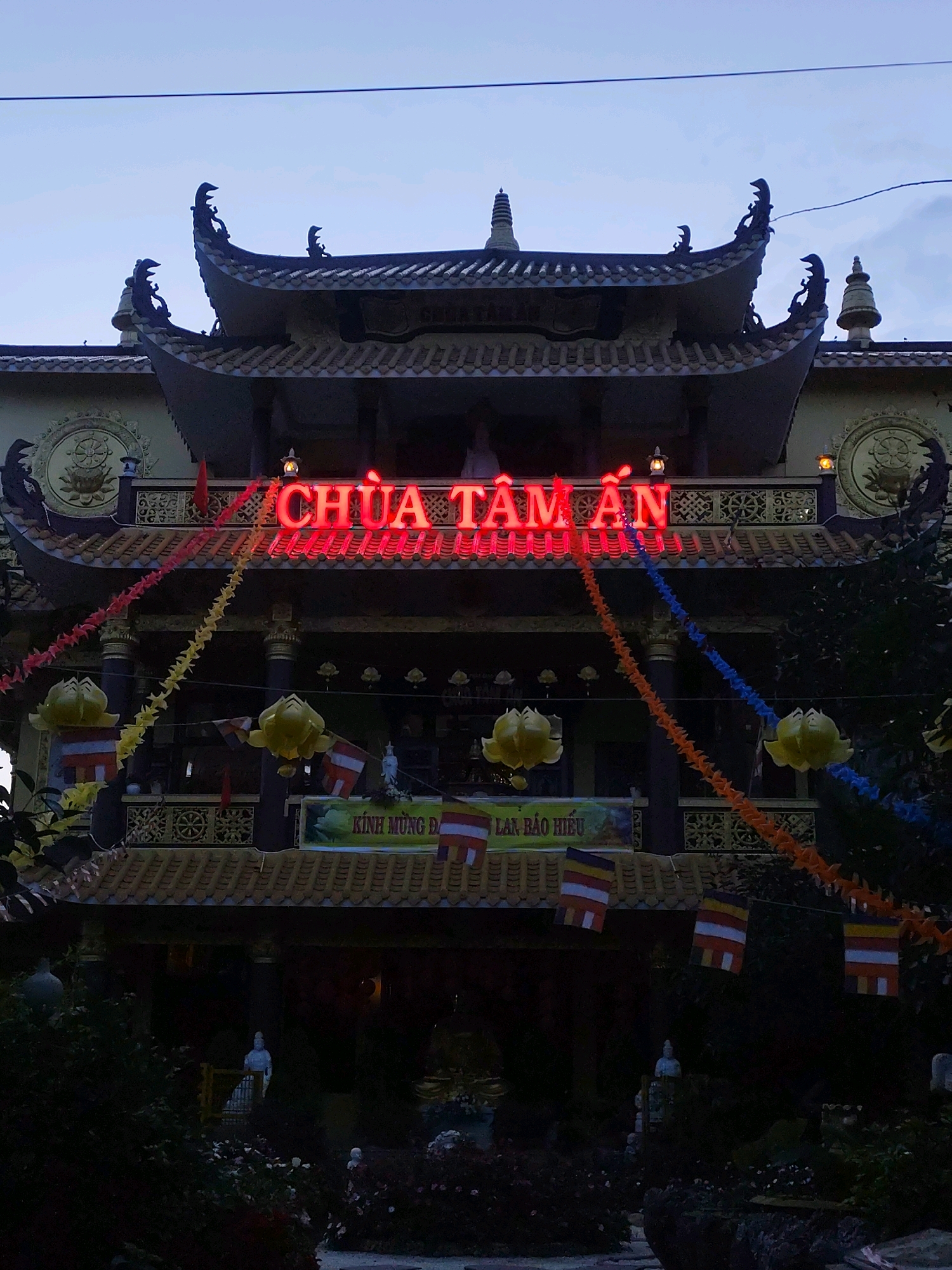 The image shows the entrance of Chua Tam An, a Buddhist temple in Da Lat, Vietnam. The temple features traditional East Asian architecture with ornate roofs and decorative elements. Red neon lights spell out "CHUA TAM AN" above the entrance, and colorful paper garlands hang across the facade. Yellow lotus-shaped lanterns are also suspended, and several small flags, some bearing the Buddhist ensign, are visible. A statue of a figure, likely a Buddhist deity, is present within the temple's entryway, partially obscured by shadows and foliage. The scene is illuminated by the temple's lights and the fading daylight, suggesting it is either dusk or dawn. The surrounding area appears to be landscaped with plants and trees.