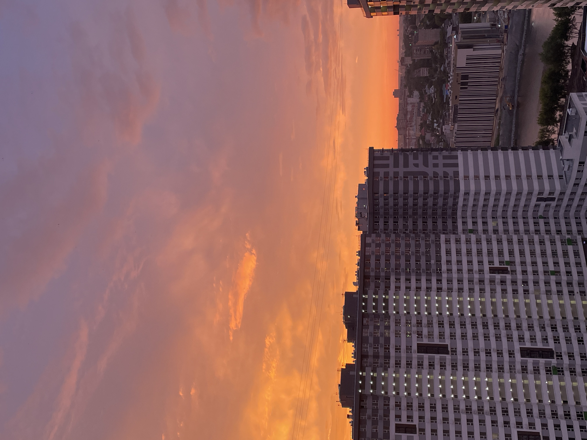 This is a landscape-oriented photograph of a city skyline at sunset. The sky is filled with a dramatic display of warm colors, ranging from deep purples and pinks on the left to vibrant oranges and yellows on the right, with clouds illuminated by the setting sun.
In the foreground, dominating the right side of the frame, are several tall, modern buildings. These buildings have a distinctive striped facade with many windows, some of which are lit from within, suggesting occupancy. The architecture appears to be residential or office complexes.
Further in the distance, a dense urban area is visible, with numerous buildings of varying heights. A river or body of water runs through the city, bordered by a road and some trees. The overall impression is one of a bustling metropolitan area transitioning from day to night.
The time of day is clearly sunset, evidenced by the rich colors in the sky and the illuminated windows in the buildings. The weather appears clear, allowing for a beautiful view of the sunset. The scene evokes a sense of calm and beauty as the day concludes, with the city lights beginning to twinkle. No text is visible in the image.